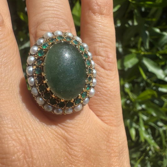 14K gold set with a large oval green Jade at its center, surrounded by concentric halos of sparkling Emeralds and delicate pearls