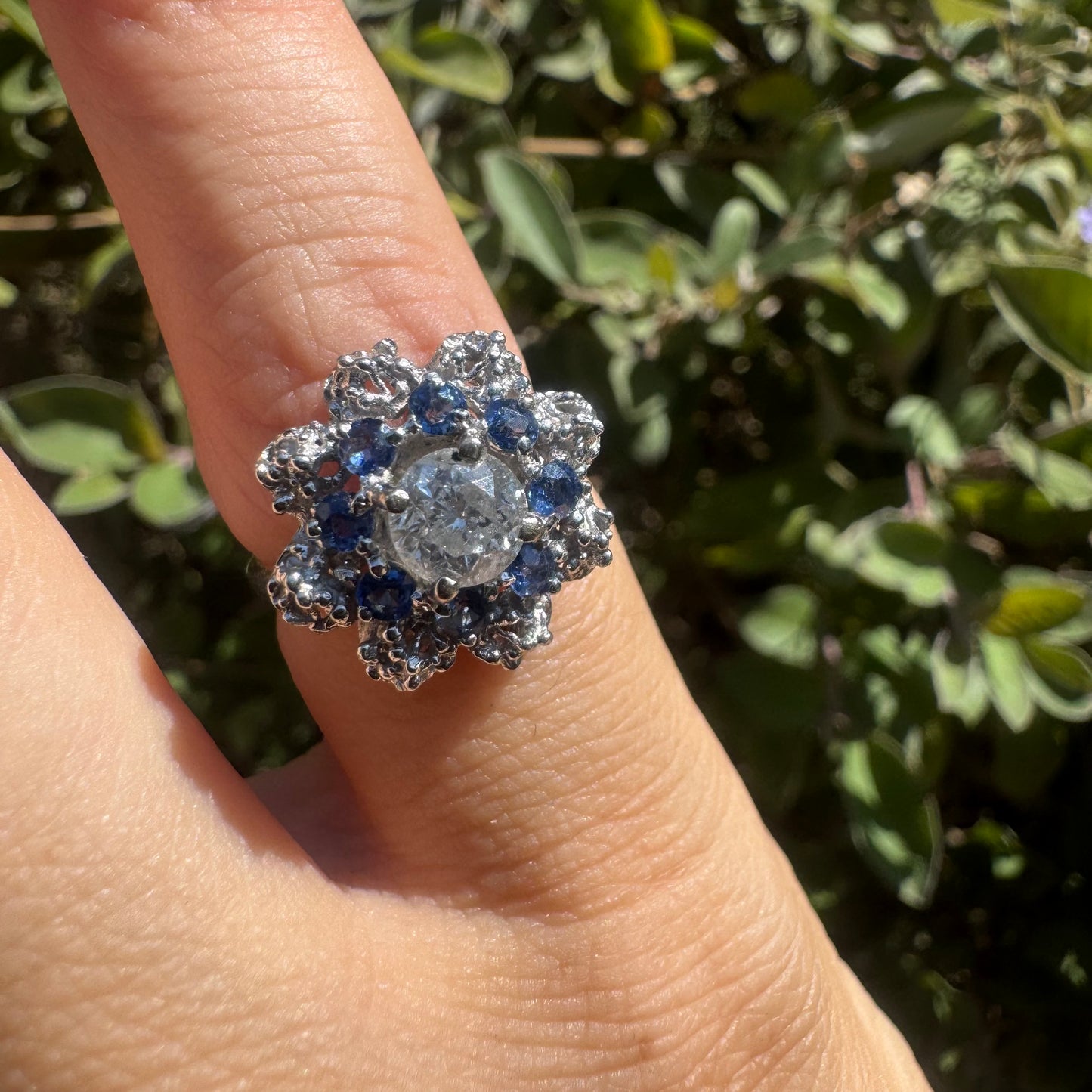 18K gold ring featuring a large round Diamond at the center, surrounded by a floral halo of purple -blue Tanzanite and & Diamonds.