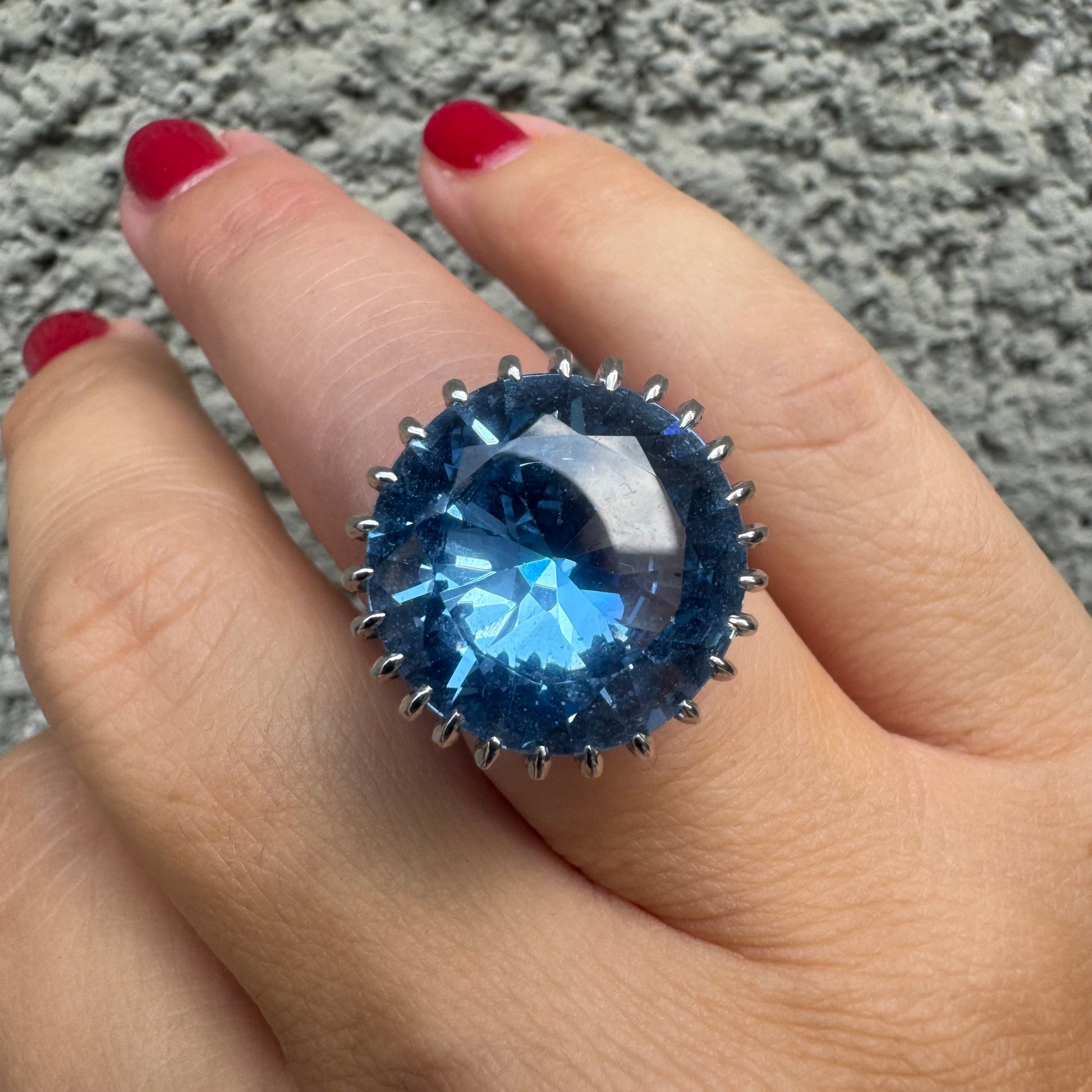 טבעת זהב לבן 14 קראט משובצת אבן טופז בצבע כחול, בולטת, בחיתוך עגול, משובצת במסגרת שיניים גבוהה ליצירת מראה מרשים ותופס עין