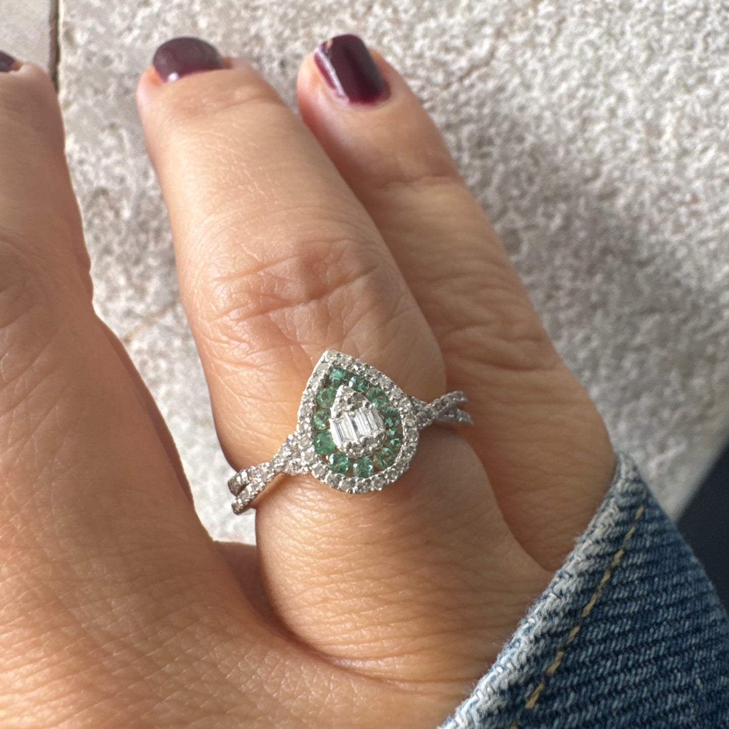 טבעת זהב צהוב 14 קראט בעיצוב טיפה (pear shape), משובצת יהלומים לבנים בשיבוץ הילה כפולה (double halo) ואבני אמרלד קטנות המקיפות מרכז ייחודי של יהלומים בחיתוך באגט