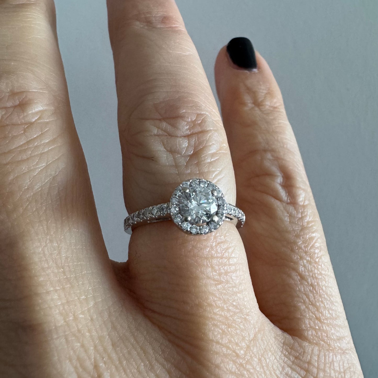 טבעת זהב לבן 14 קראט משובצת יהלום מרכזי עגול והילה נוצצת של יהלומים קטנים, עם יהלומים נוספים על החישוק.