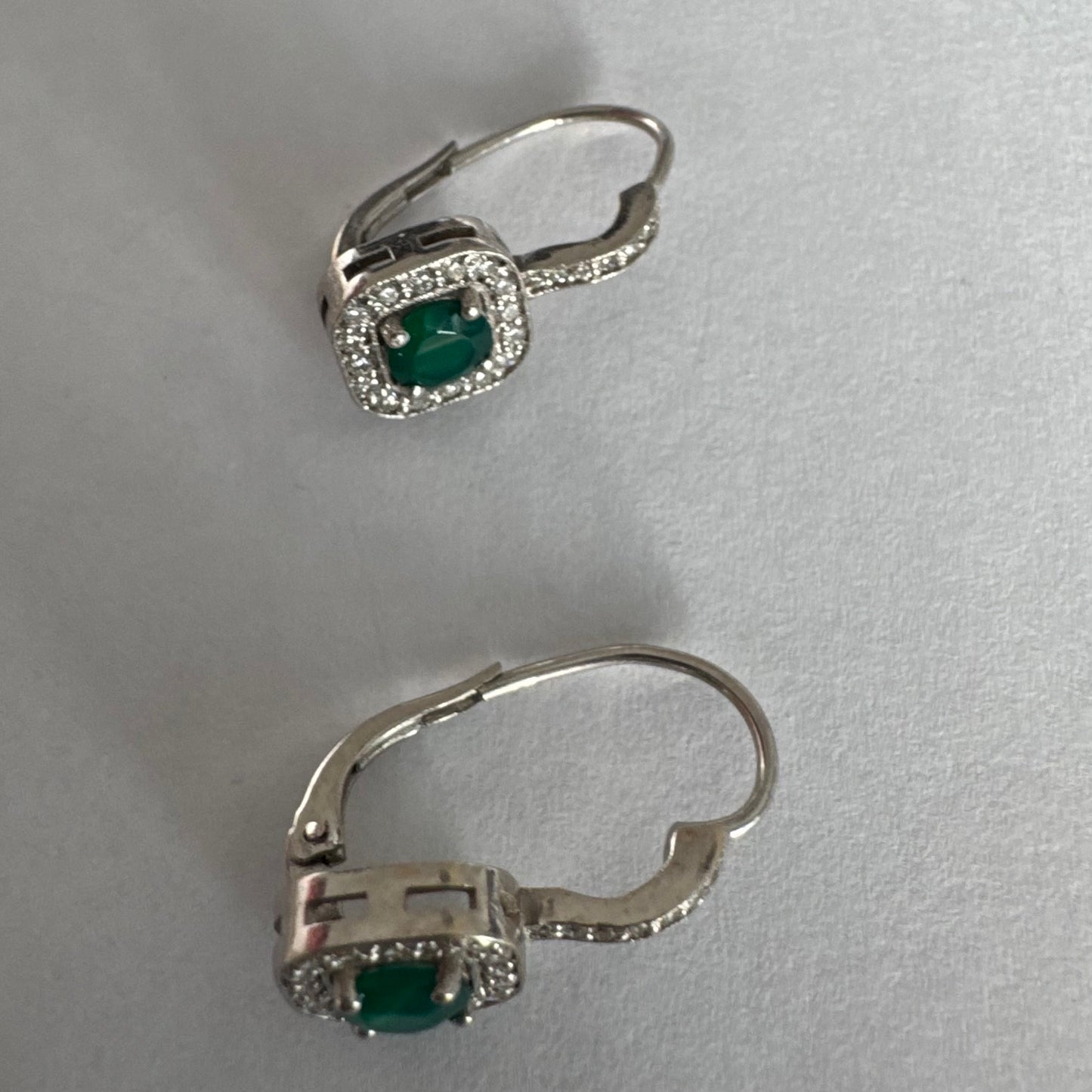 עגילי זהב לבן 14 קראט בעיצוב אלגנטי ונקי, משובצים אבן אמרלד מרכזית במראה עשיר ועמוק, מוקפת הילה מנצנצת של יהלומים.