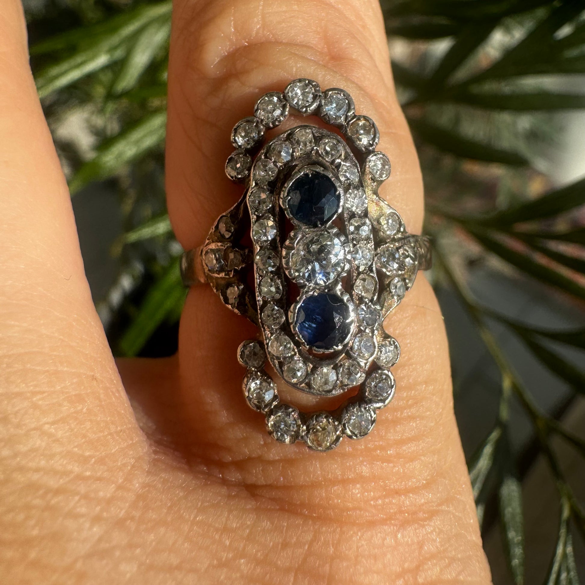 טבעת זהב לבן 14 קראט עתיקה עוצרת נשימה, בעיצוב מאורך ואלגנטי, המשובצת בשלישיית אבנים — שתי אבני ספיר בצבע כחול עמוק ובאמצע משובץ יהלום מרכזי בחיתוך עתיק.