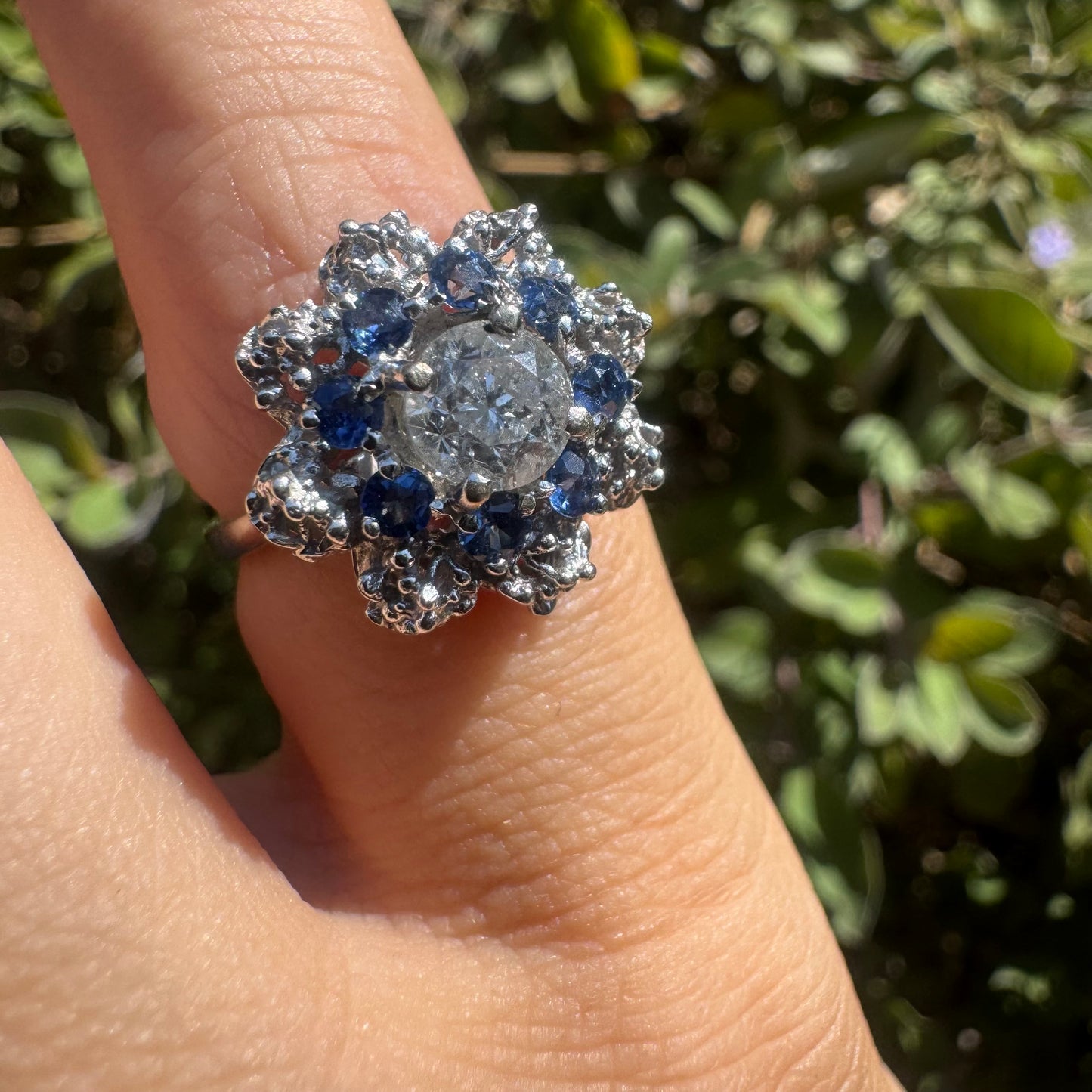 טבעת זהב לבן 18 קראט משובצת יהלום עגול גדול במרכז, מוקף בהילה בצורת פרח משובצת אבני טנזנייט בצבע כחול-סגול ויהלומים