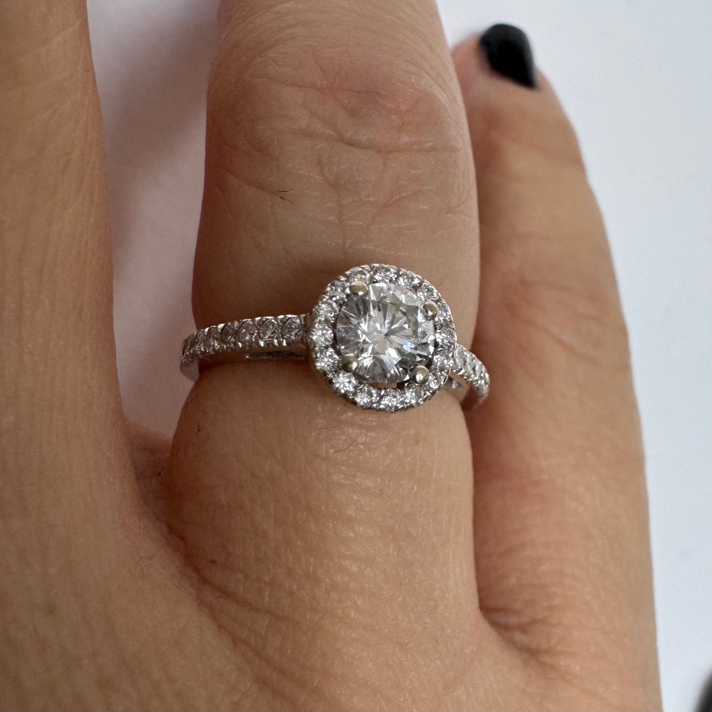 טבעת זהב לבן 14 קראט משובצת יהלום מרכזי עגול והילה נוצצת של יהלומים קטנים, עם יהלומים נוספים על החישוק.