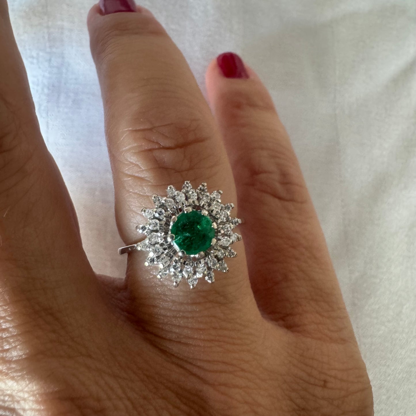 טבעת זהב לבן 14 קראט, משובצת אבן אמרלד ירוקה במרכז, מוקפת הילה עשירה של יהלומים בעיצוב קלאסי ואלגנטי. 