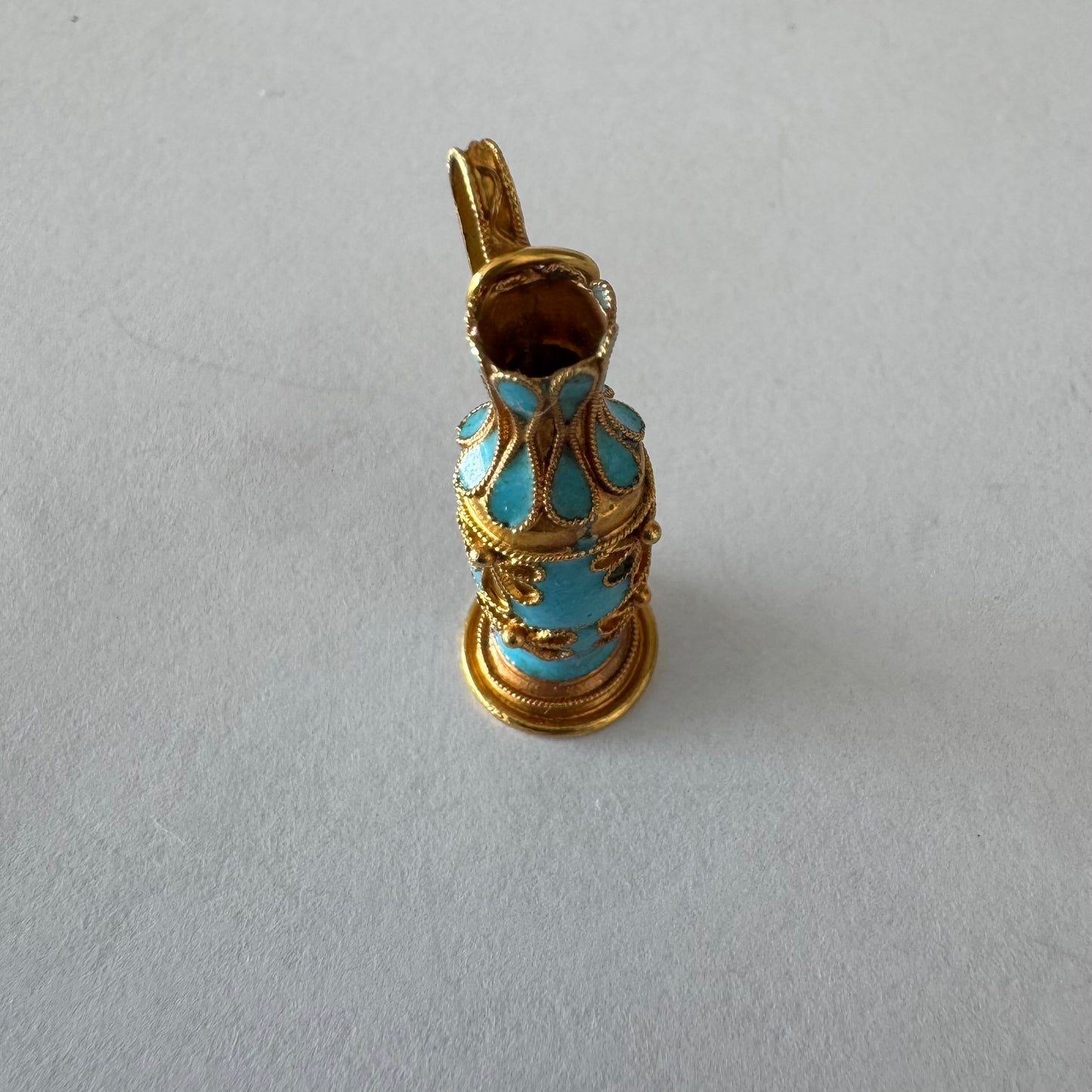 תליון זהב 18 קראט ייחודי בצורת כד מיניאטורי, מעוטר באמייל בגוון טורקיז ובעבודת פיליגרן מוזהבת עשירה בדוגמאות פרחוניות.