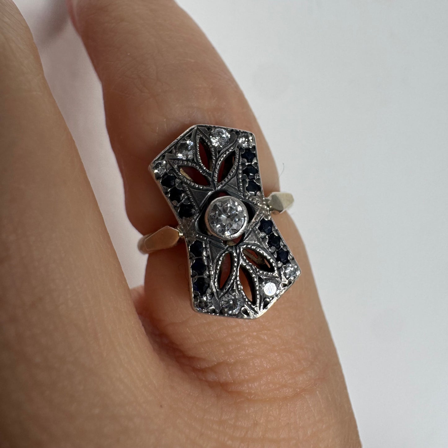 טבעת זהב 14 קראט בעיצוב אר-דקו גיאומטרי, משובצת יהלום מרכזי ומסגרת של אבני ספיר כהות ויהלומים נוצצים.