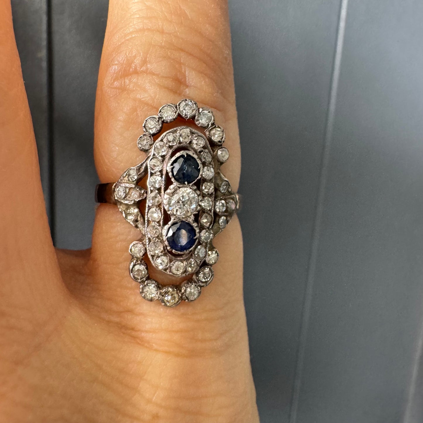 טבעת זהב לבן 14 קראט עתיקה עוצרת נשימה, בעיצוב מאורך ואלגנטי, המשובצת בשלישיית אבנים — שתי אבני ספיר בצבע כחול עמוק ובאמצע משובץ יהלום מרכזי בחיתוך עתיק.