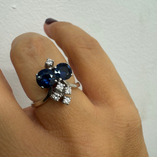 טבעת זהב לבן 14 קראט משובצת שתי אבני ספיר בגוון כחול עמוק ובאשכולות יהלומים לבנים מנצנצים.