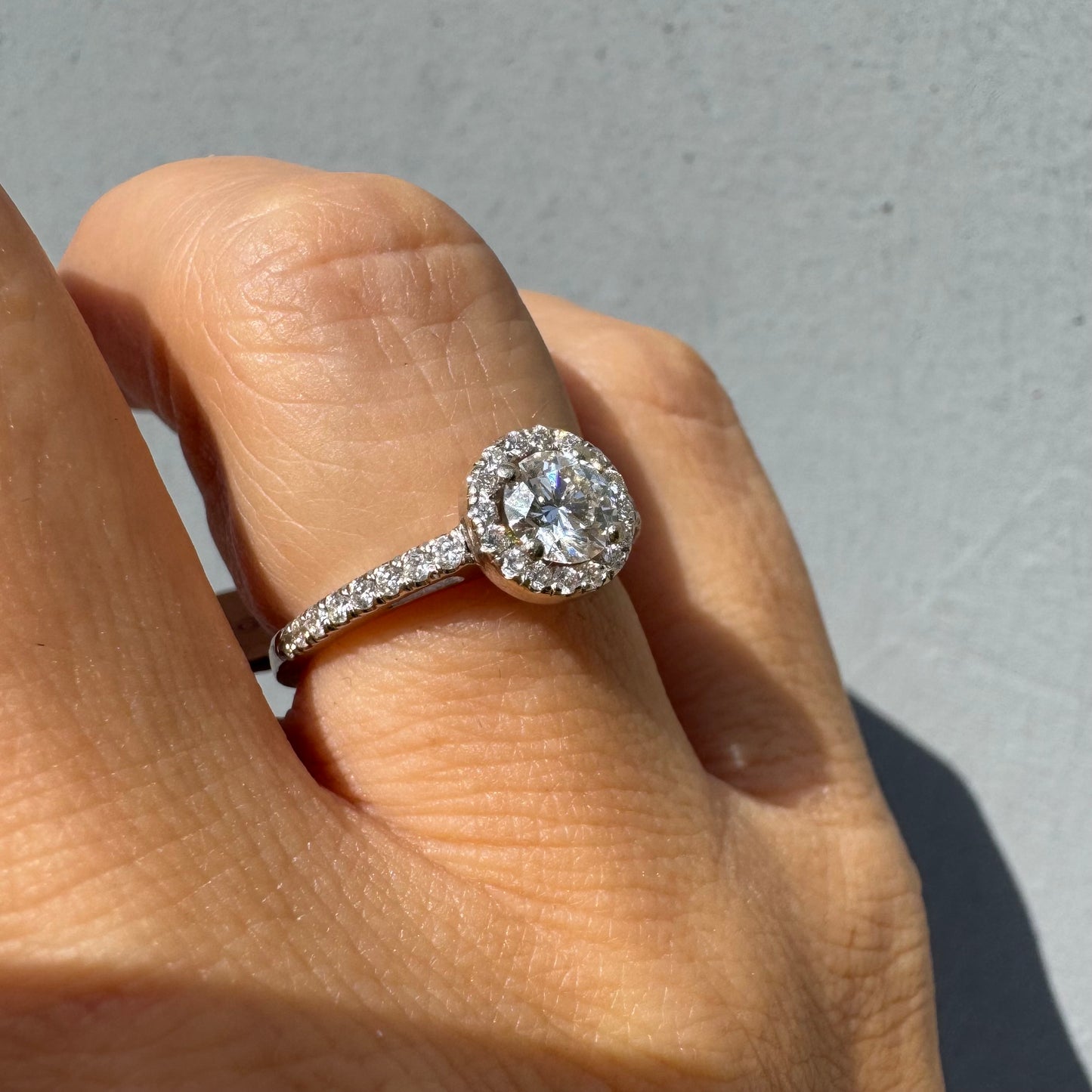 טבעת זהב לבן 14 קראט משובצת יהלום מרכזי עגול והילה נוצצת של יהלומים קטנים, עם יהלומים נוספים על החישוק.