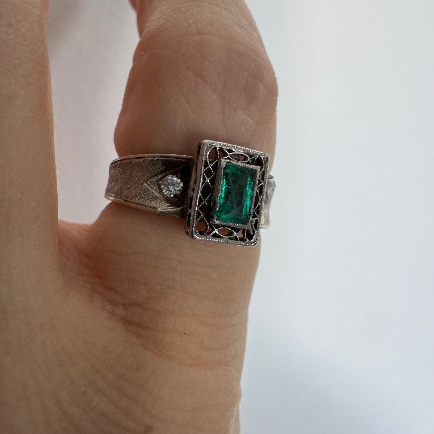 טבעת זהב לבן 14 קראט משובצת אבן אמרלד בחיתוך מלבני (Emerald cut) במרכז, מוקפת במסגרת מלבנית עם עבודת פיליגרן עדינה.