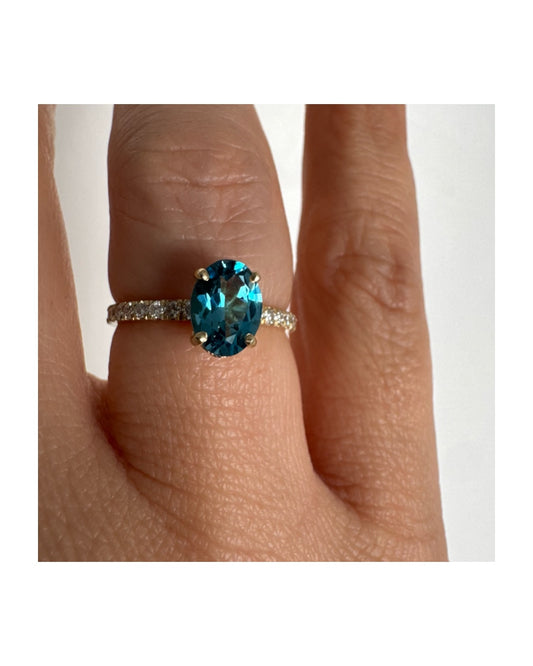 טבעת זהב צהוב 14 קראט משובצת אבן טופז כחולה אובלית בגוון טורקיז־כחול עמוק, באחיזה של ארבע שיניים עדינות ויהלומים.