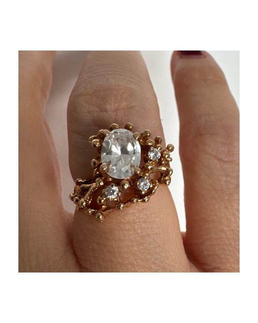 טבעת זהב צהוב 14 קראט משובצת אבן מרכזית אובלית שקופה, וסביבה אבנים שקופות נוספות בעיצוב פתוח ואורגני.