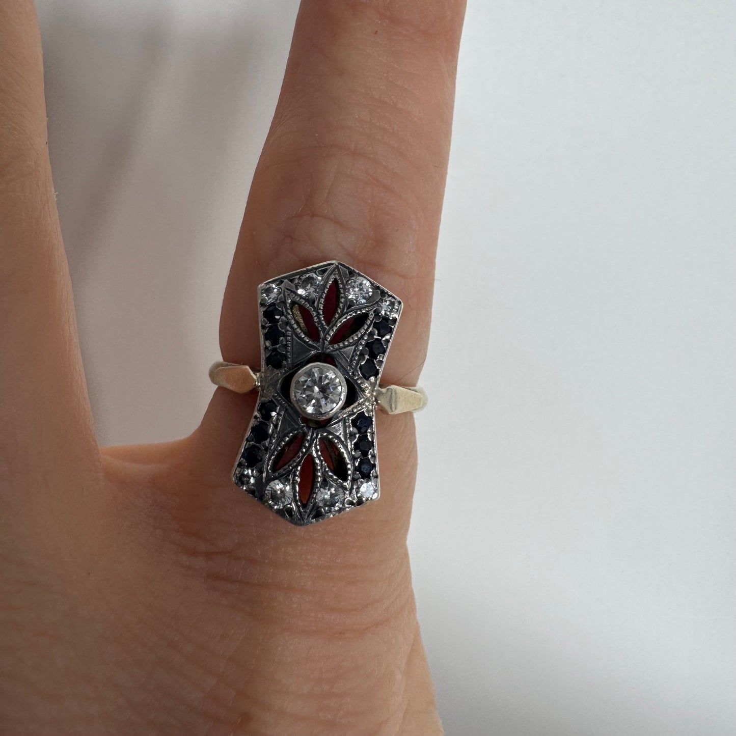 טבעת זהב 14 קראט בעיצוב אר-דקו גיאומטרי, משובצת יהלום מרכזי ומסגרת של אבני ספיר כהות ויהלומים נוצצים.