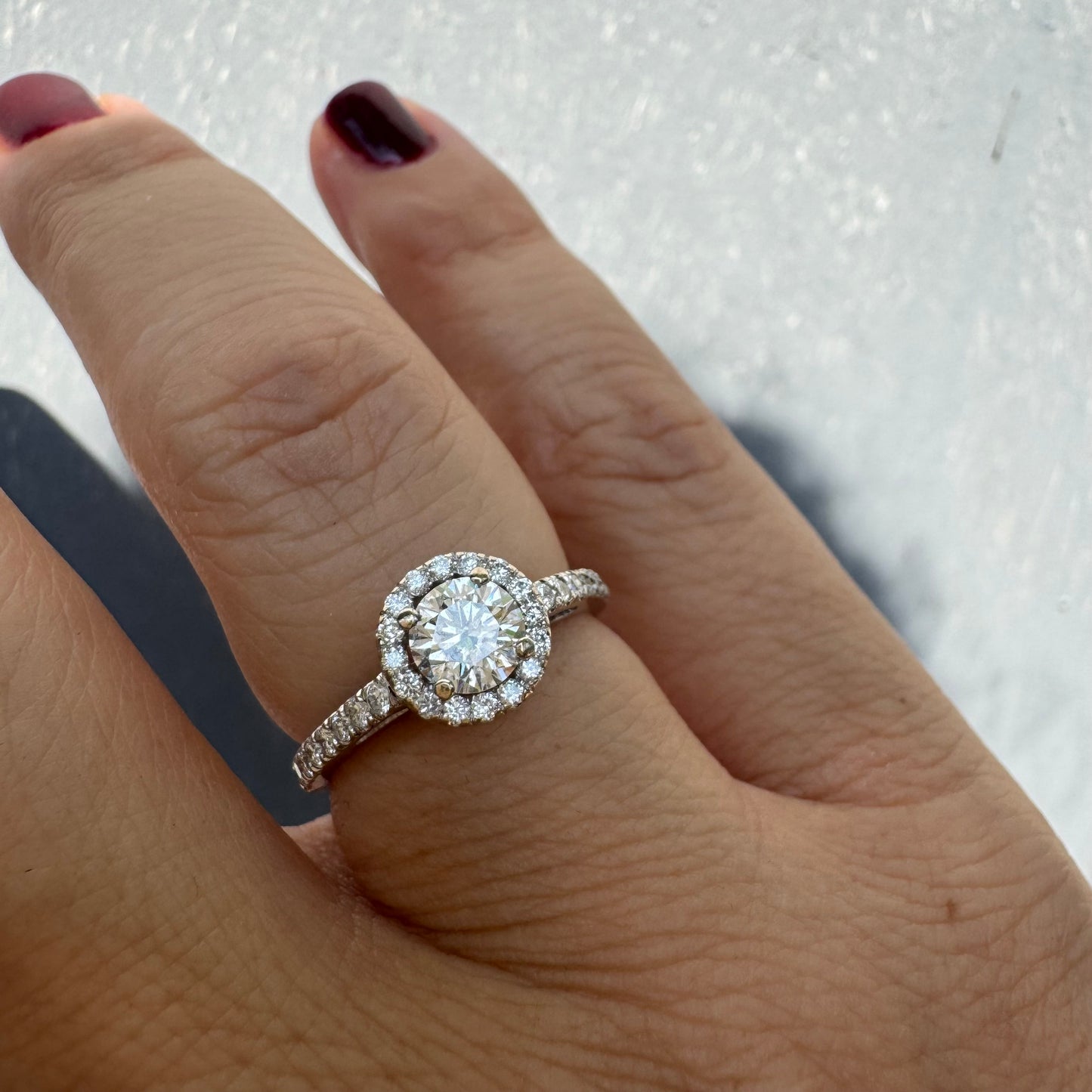 טבעת זהב לבן 14 קראט משובצת יהלום מרכזי עגול והילה נוצצת של יהלומים קטנים, עם יהלומים נוספים על החישוק.