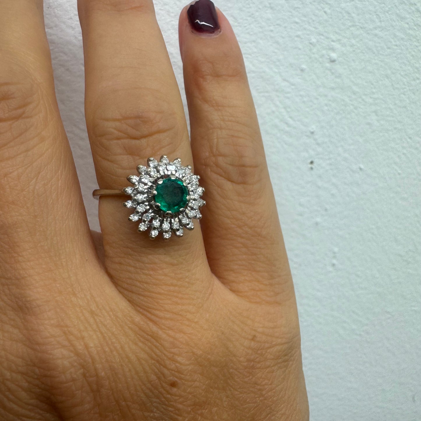 טבעת זהב לבן 14 קראט, משובצת אבן אמרלד ירוקה במרכז, מוקפת הילה עשירה של יהלומים בעיצוב קלאסי ואלגנטי. 