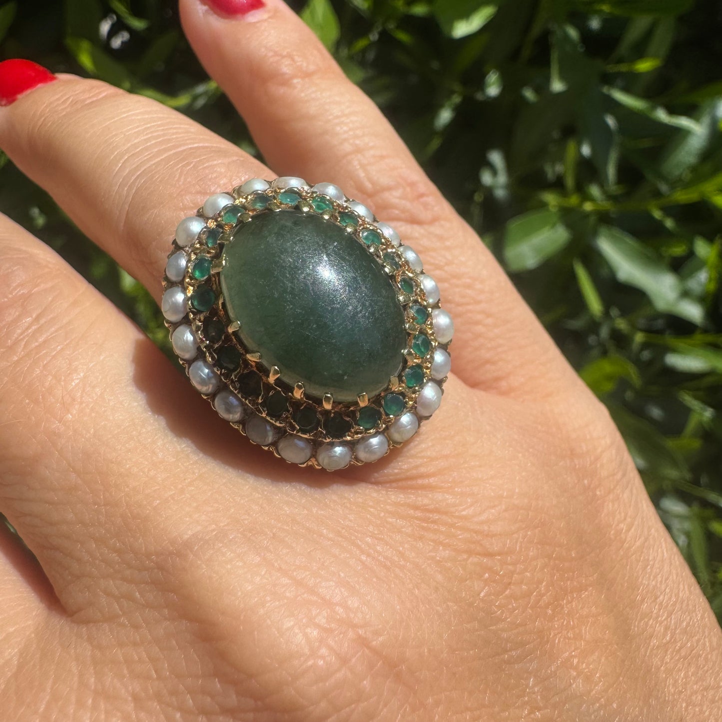 14K gold set with a large oval green Jade at its center, surrounded by concentric halos of sparkling Emeralds and delicate pearls