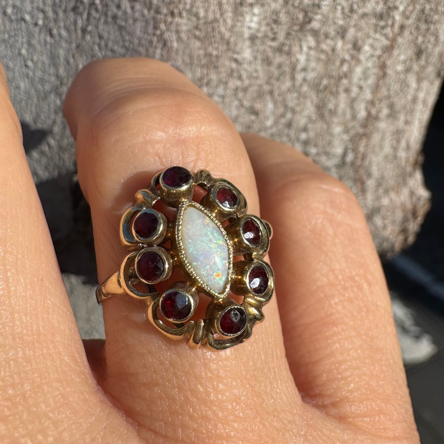 טבעת זהב צהוב 14 קראט משובצת באבן אופל מרכזית בחיתוך מרקיזה, מוקפת באבני גרנט עגולות כהות.