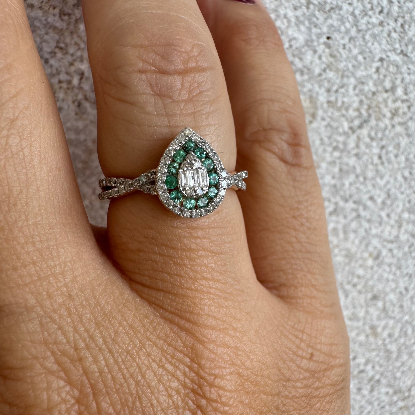 טבעת זהב צהוב 14 קראט בעיצוב טיפה (pear shape), משובצת יהלומים לבנים בשיבוץ הילה כפולה (double halo) ואבני אמרלד קטנות המקיפות מרכז ייחודי של יהלומים בחיתוך באגט