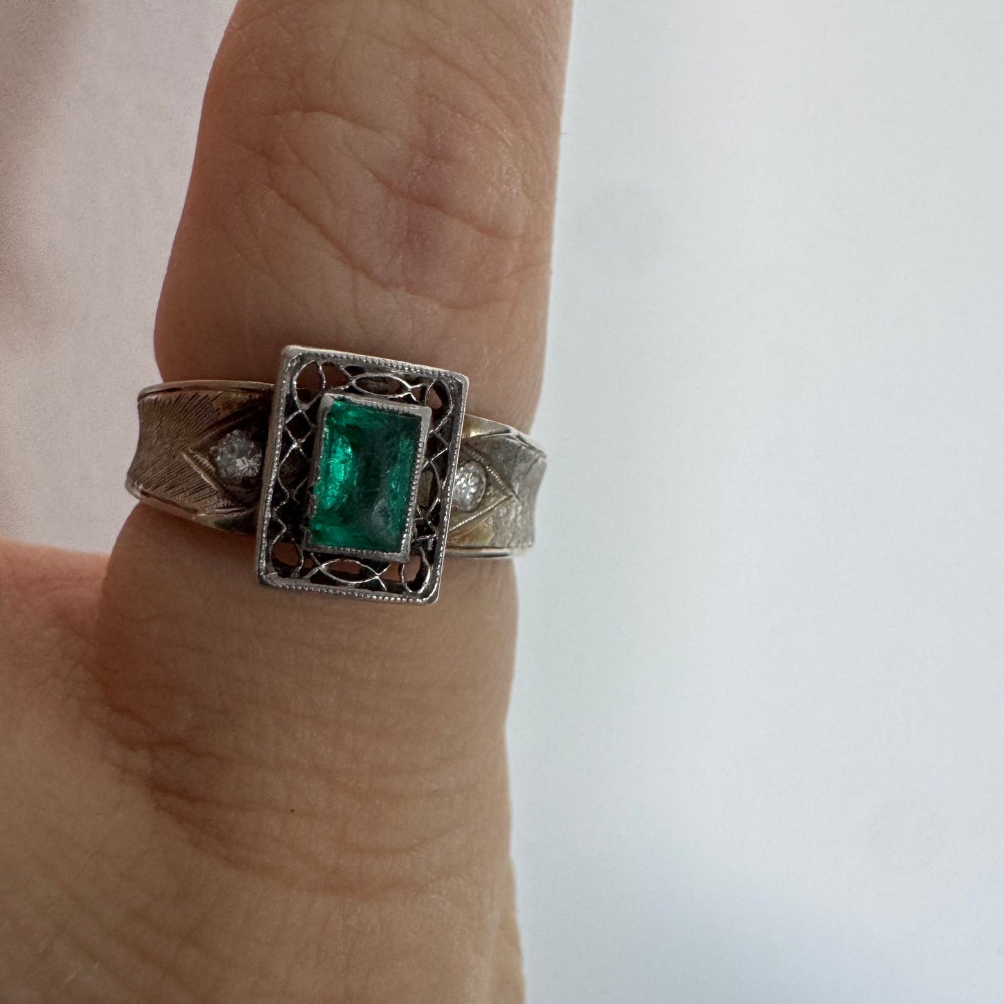 טבעת זהב לבן 14 קראט משובצת אבן אמרלד בחיתוך מלבני (Emerald cut) במרכז, מוקפת במסגרת מלבנית עם עבודת פיליגרן עדינה.