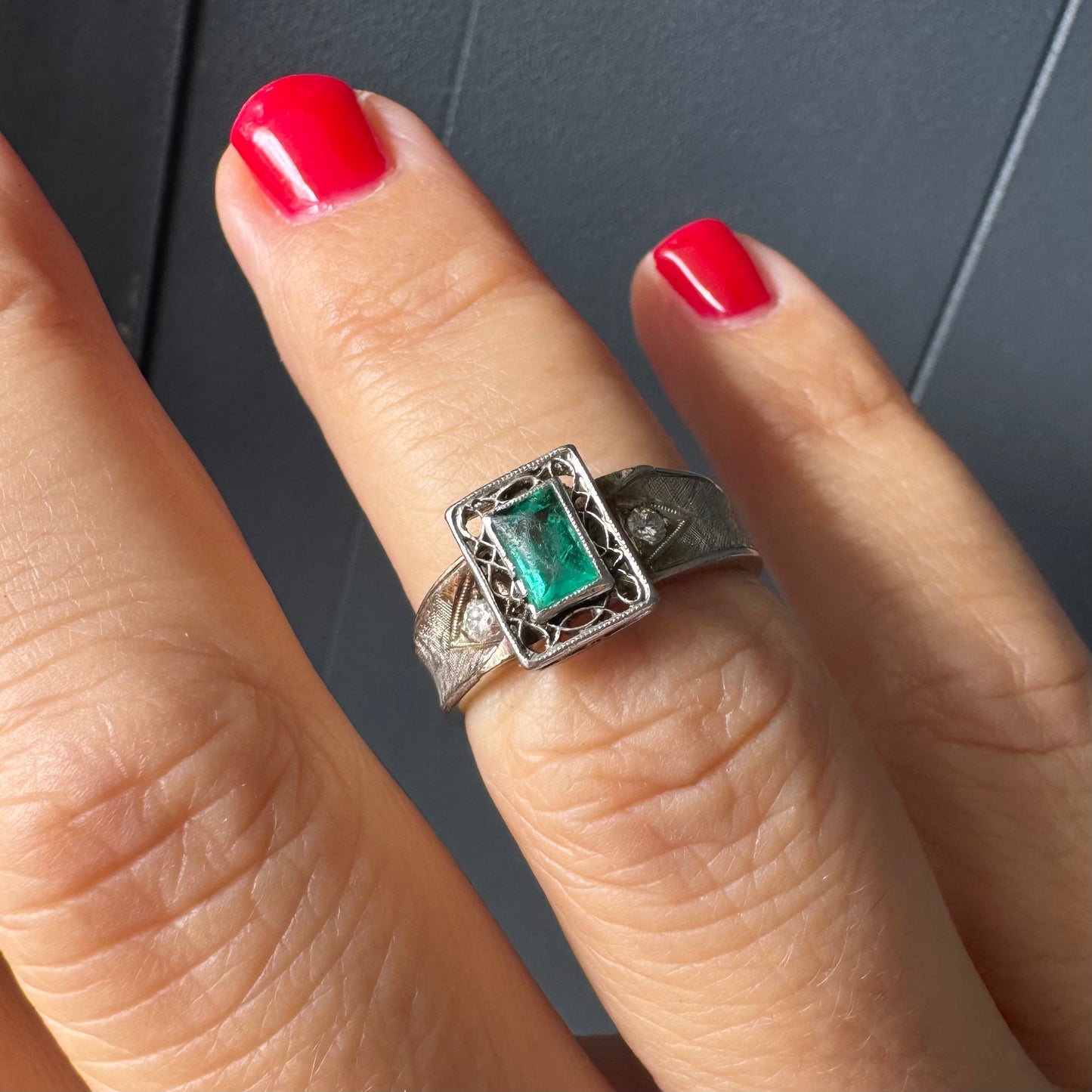 טבעת זהב לבן 14 קראט משובצת אבן אמרלד בחיתוך מלבני (Emerald cut) במרכז, מוקפת במסגרת מלבנית עם עבודת פיליגרן עדינה.