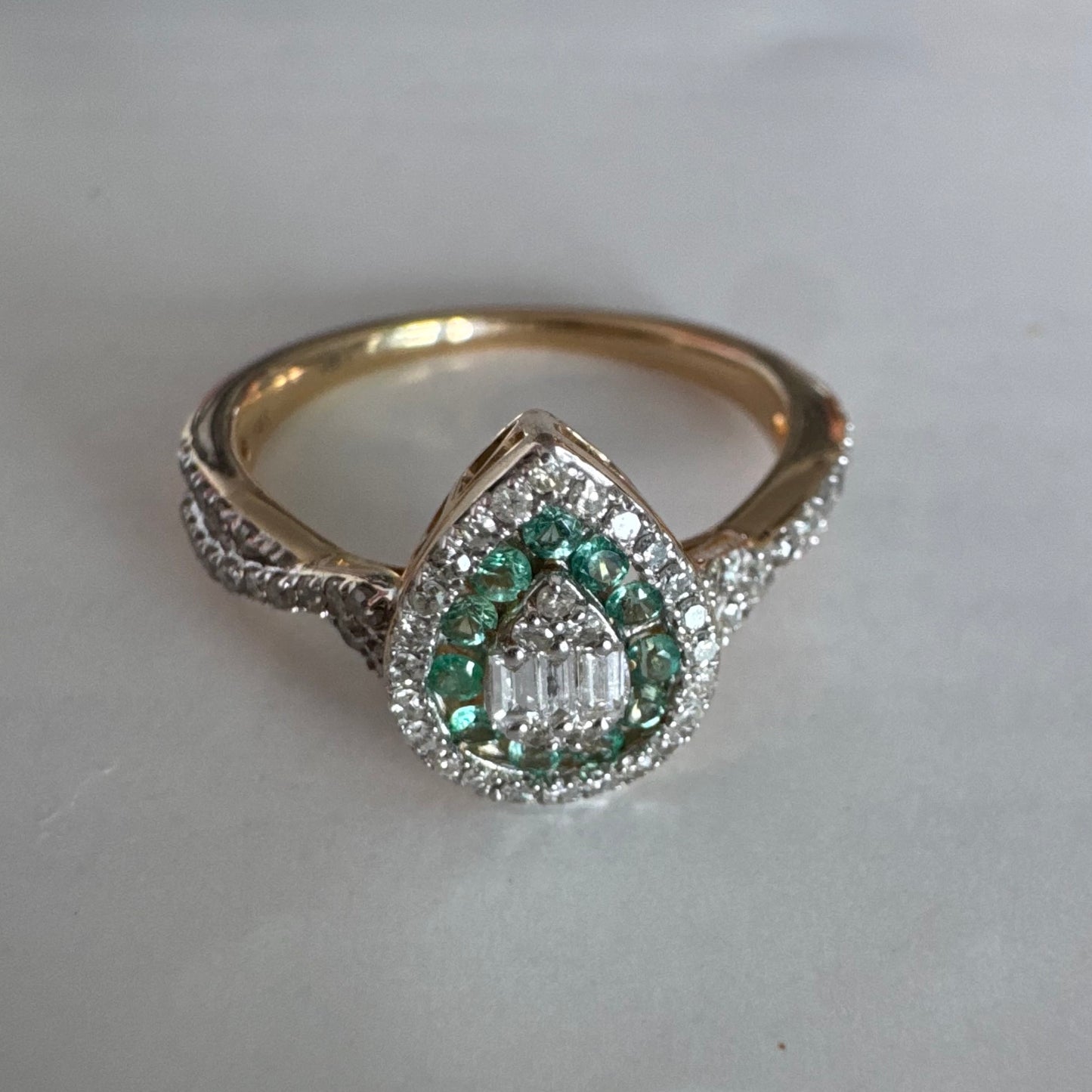 טבעת זהב צהוב 14 קראט בעיצוב טיפה (pear shape), משובצת יהלומים לבנים בשיבוץ הילה כפולה (double halo) ואבני אמרלד קטנות המקיפות מרכז ייחודי של יהלומים בחיתוך באגט