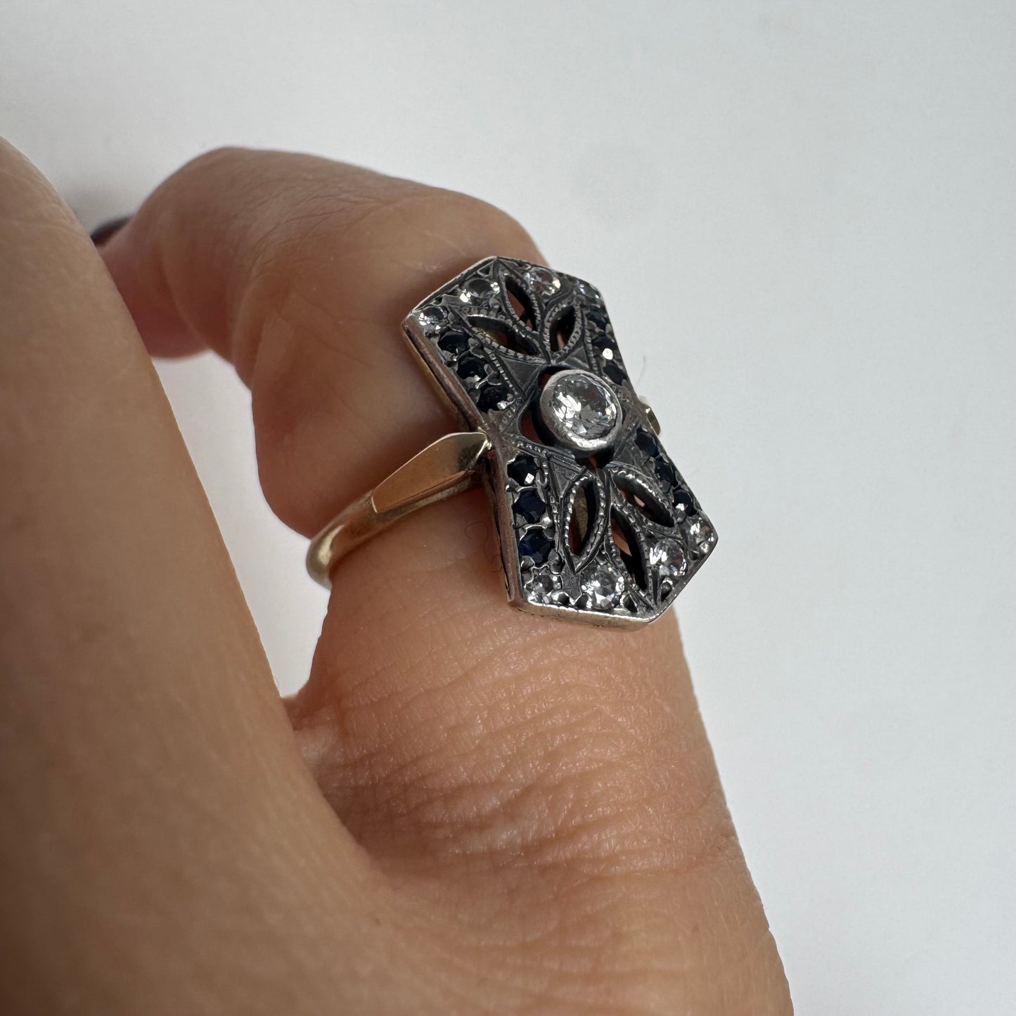 טבעת זהב 14 קראט בעיצוב אר-דקו גיאומטרי, משובצת יהלום מרכזי ומסגרת של אבני ספיר כהות ויהלומים נוצצים.