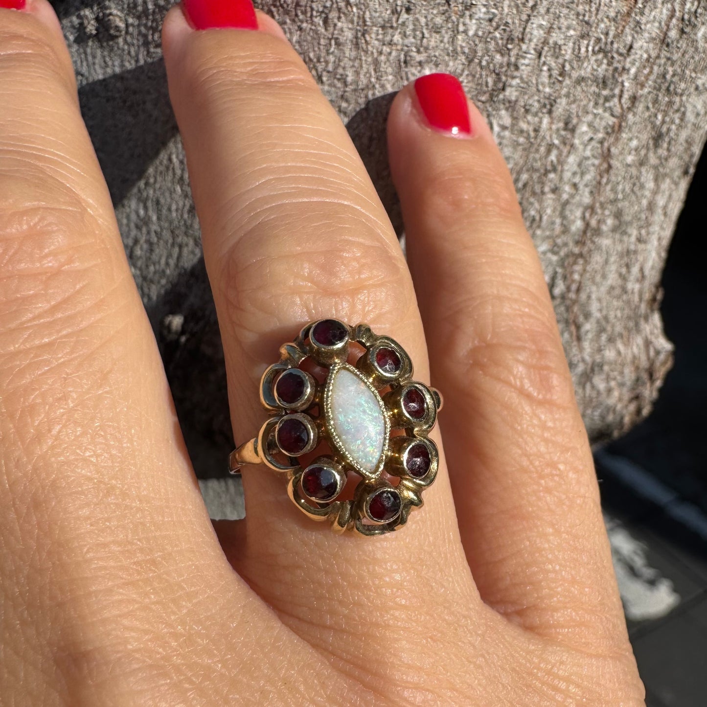 טבעת זהב צהוב 14 קראט משובצת באבן אופל מרכזית בחיתוך מרקיזה, מוקפת באבני גרנט עגולות כהות.