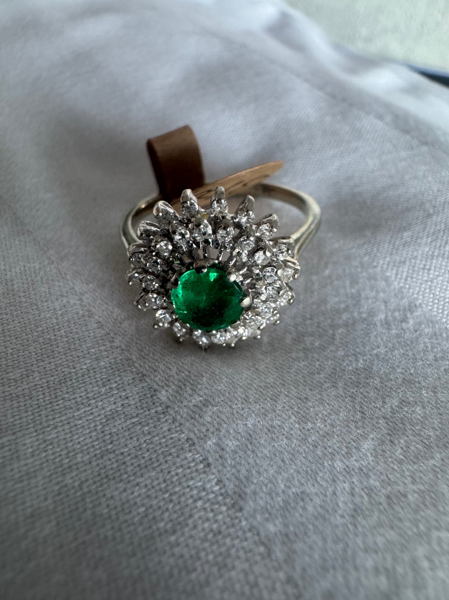 טבעת זהב לבן 14 קראט, משובצת אבן אמרלד ירוקה במרכז, מוקפת הילה עשירה של יהלומים בעיצוב קלאסי ואלגנטי. 