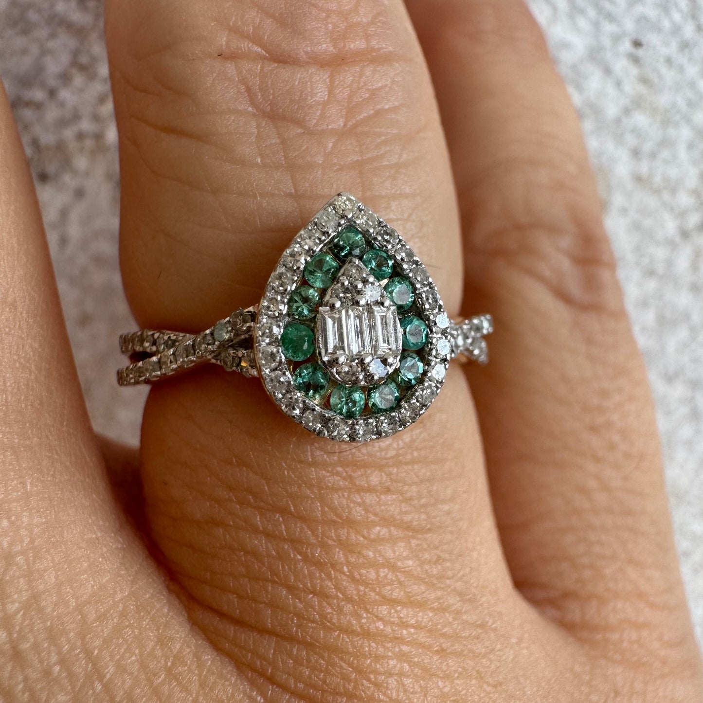 טבעת זהב צהוב 14 קראט בעיצוב טיפה (pear shape), משובצת יהלומים לבנים בשיבוץ הילה כפולה (double halo) ואבני אמרלד קטנות המקיפות מרכז ייחודי של יהלומים בחיתוך באגט