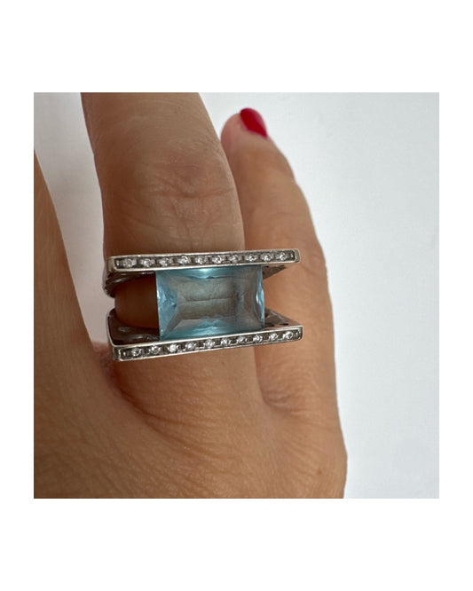 טבעת זהב לבן 18 קראט בעיצוב מודרני ונועז, משובצת אבן אקוומרין מרהיבה במרכז, הנתמכת בין שני פסי זהב משובצים יהלומים עדינים.