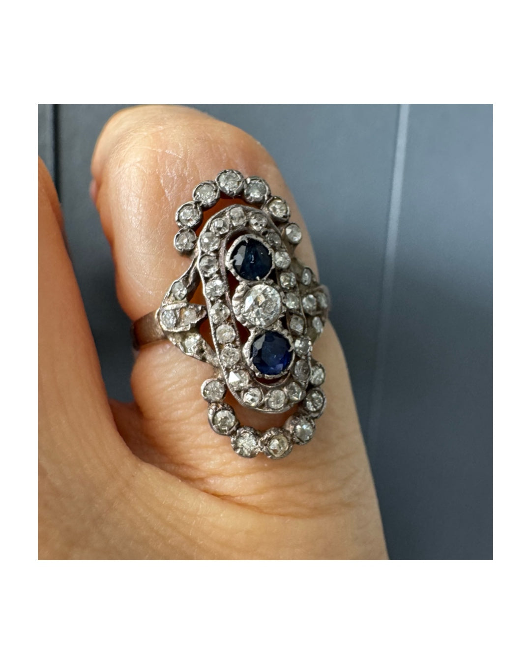 טבעת זהב לבן 14 קראט עתיקה עוצרת נשימה, בעיצוב מאורך ואלגנטי, המשובצת בשלישיית אבנים — שתי אבני ספיר בצבע כחול עמוק ובאמצע משובץ יהלום מרכזי בחיתוך עתיק.