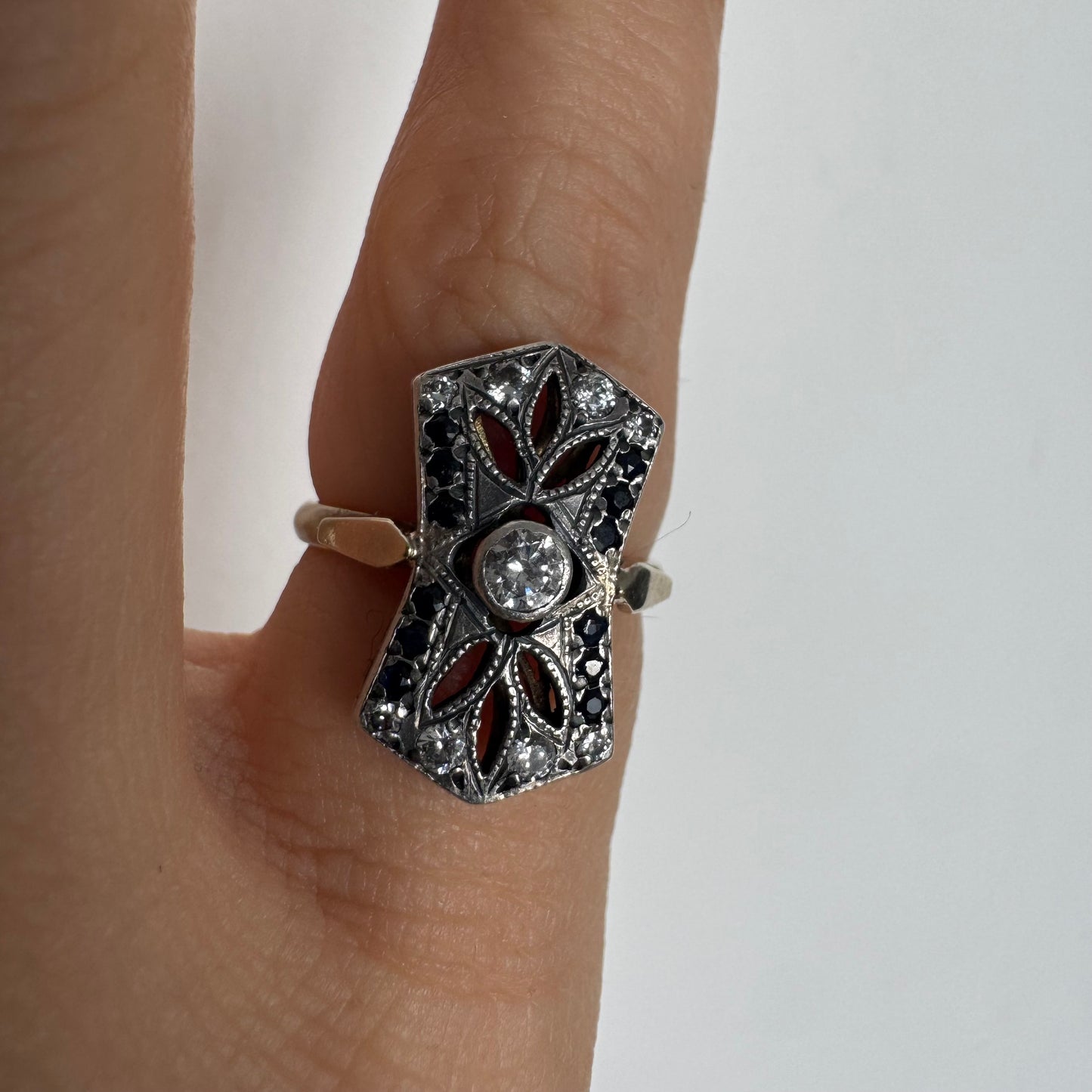 טבעת זהב 14 קראט בעיצוב אר-דקו גיאומטרי, משובצת יהלום מרכזי ומסגרת של אבני ספיר כהות ויהלומים נוצצים.