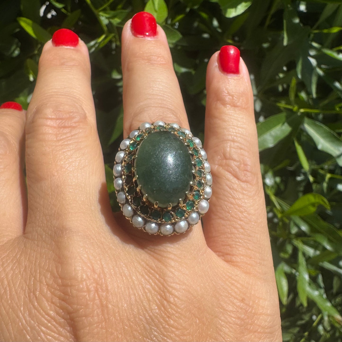 14K gold set with a large oval green Jade at its center, surrounded by concentric halos of sparkling Emeralds and delicate pearls