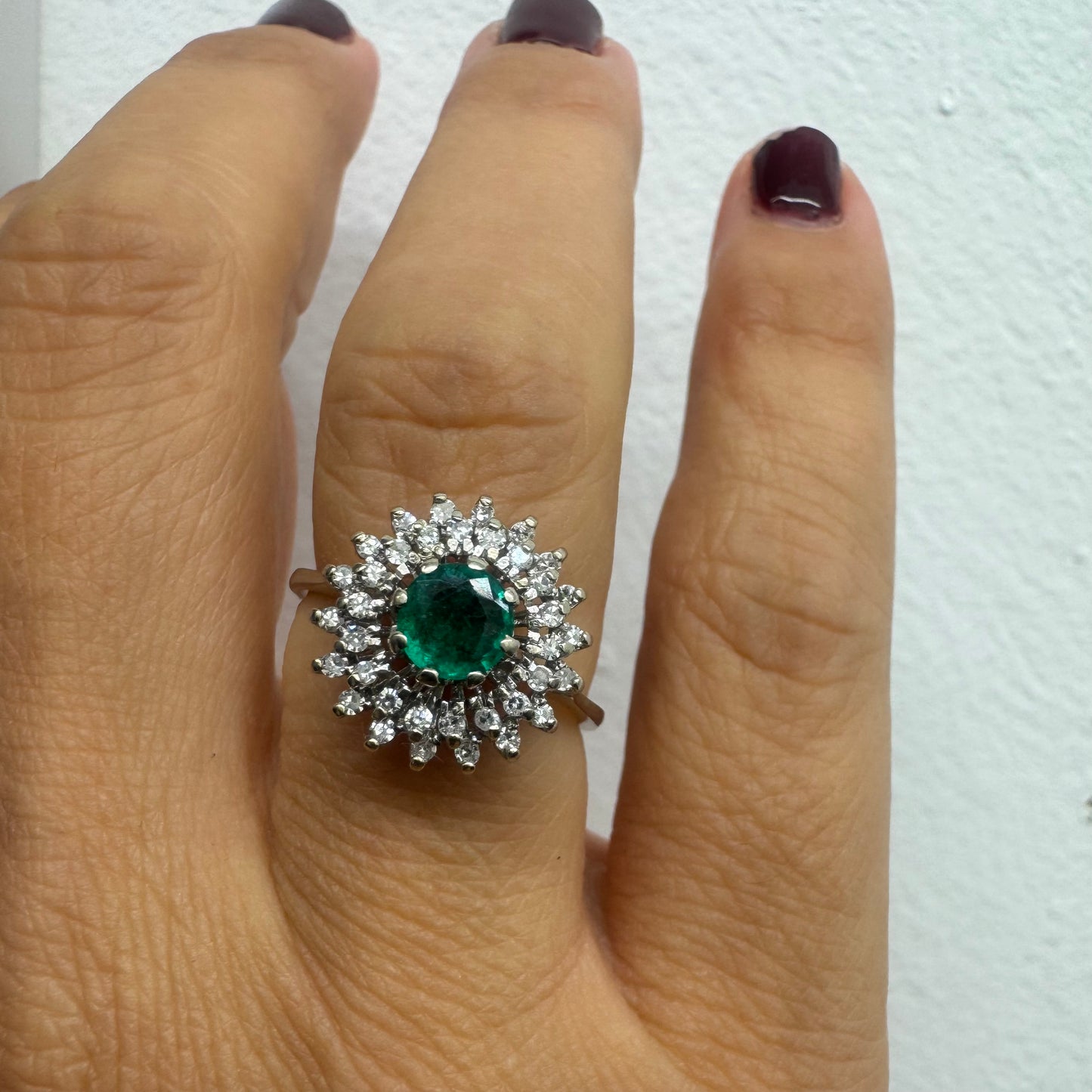 טבעת זהב לבן 14 קראט, משובצת אבן אמרלד ירוקה במרכז, מוקפת הילה עשירה של יהלומים בעיצוב קלאסי ואלגנטי. 