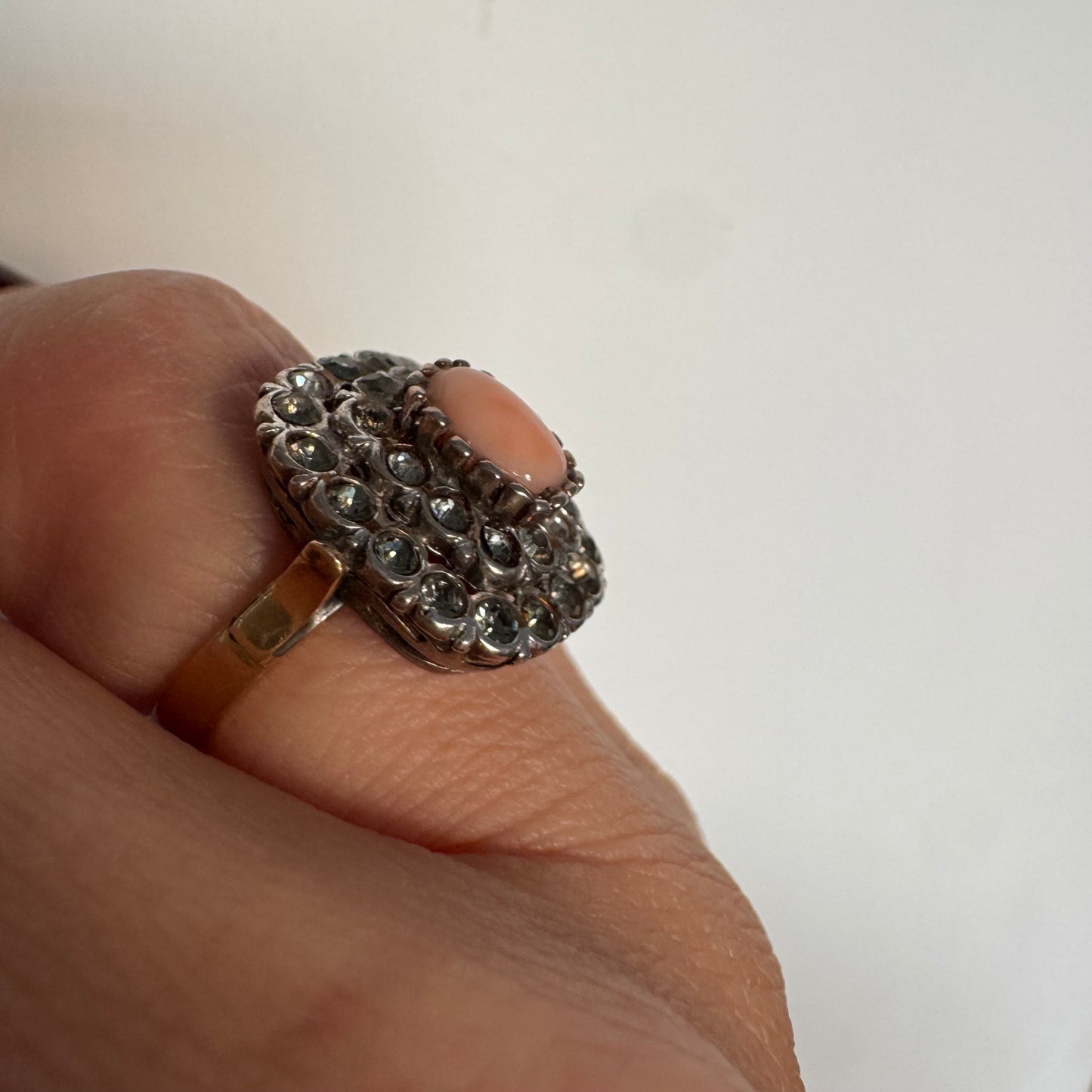 טבעת וינטג׳ עם חלק תחתון מזהב 14 קראט וחלק עליון עשוי כסף משובצת אבן קורל בהירה במרכז ומסביבה אבנים בהירות שאינן יהלומים.