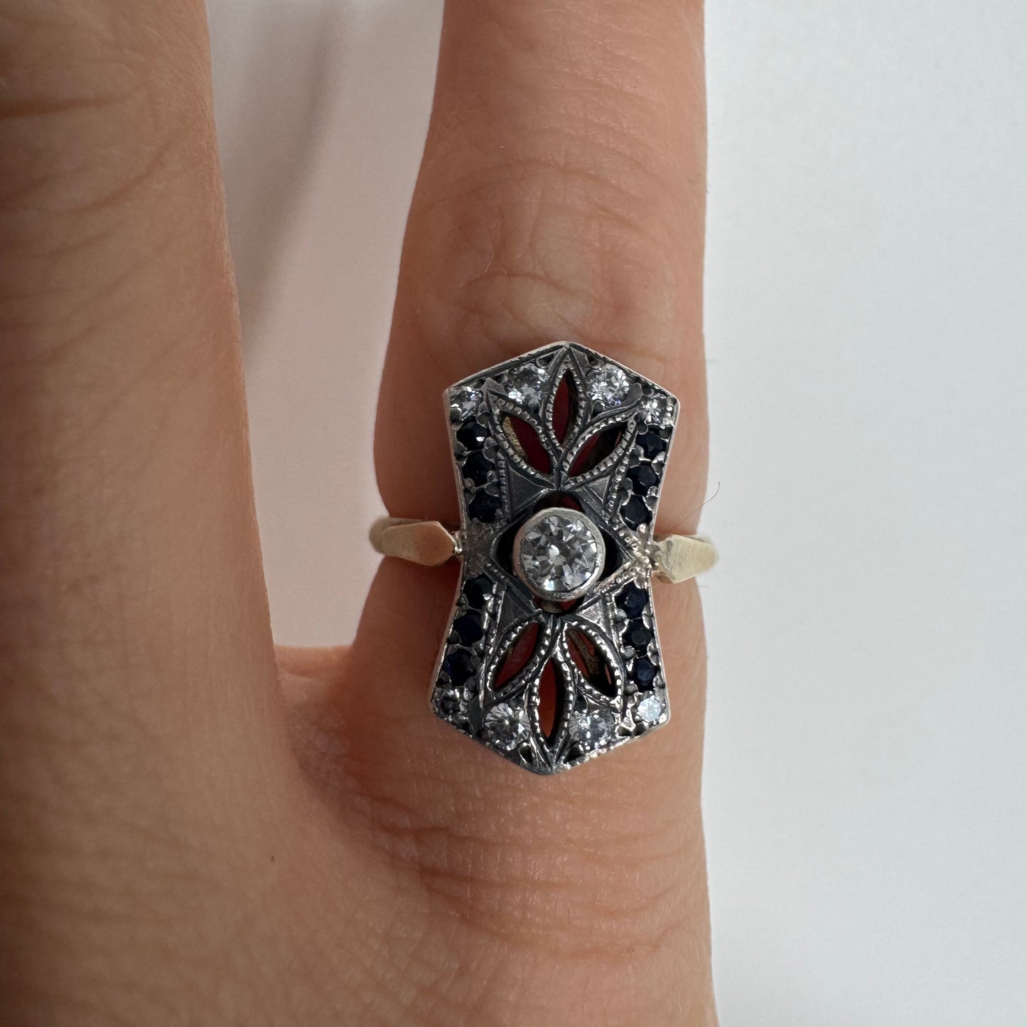 טבעת זהב 14 קראט בעיצוב אר-דקו גיאומטרי, משובצת יהלום מרכזי ומסגרת של אבני ספיר כהות ויהלומים נוצצים.