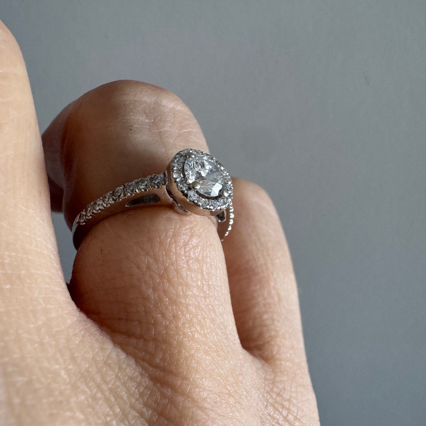 טבעת זהב לבן 14 קראט משובצת יהלום מרכזי עגול והילה נוצצת של יהלומים קטנים, עם יהלומים נוספים על החישוק.