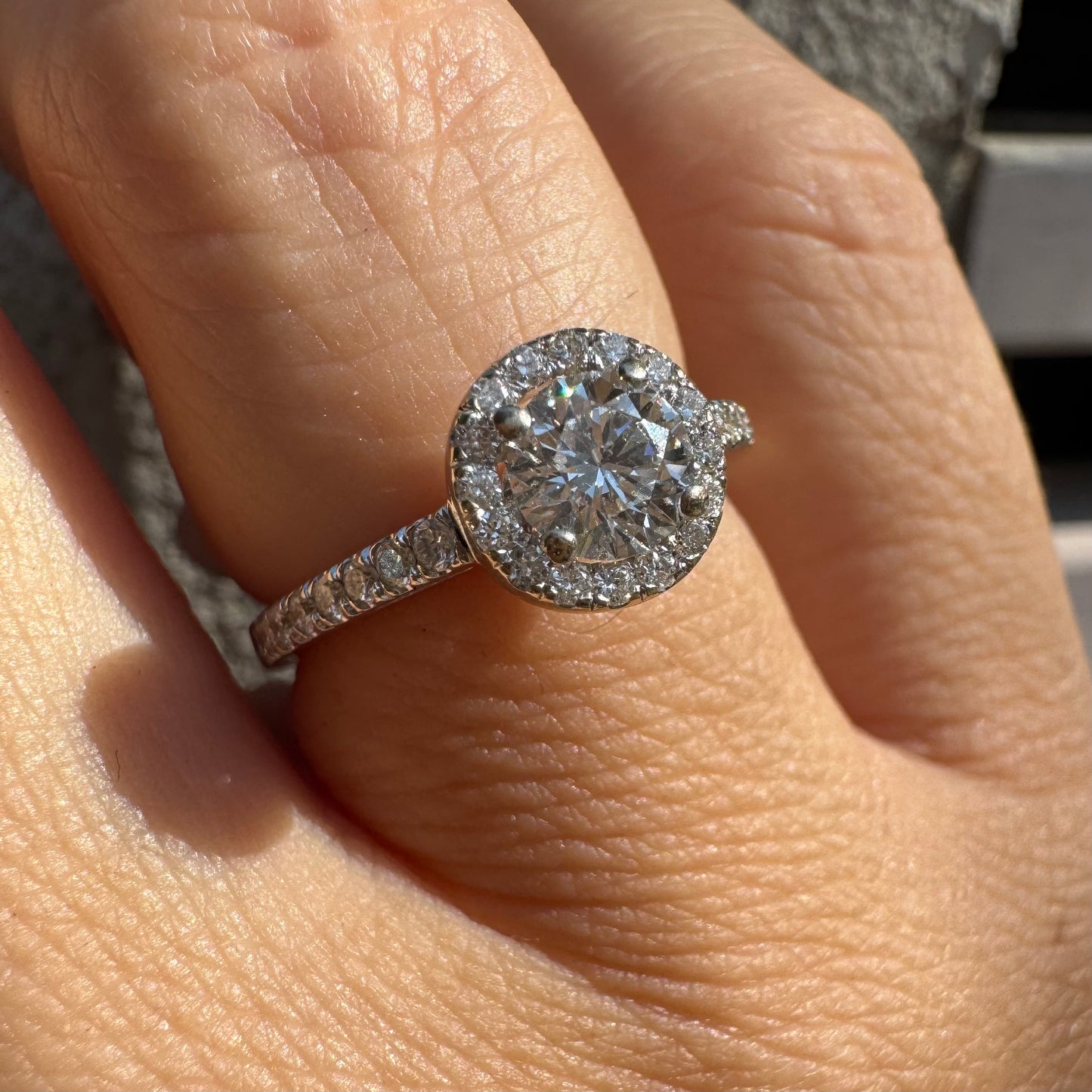 טבעת זהב לבן 14 קראט משובצת יהלום מרכזי עגול והילה נוצצת של יהלומים קטנים, עם יהלומים נוספים על החישוק.