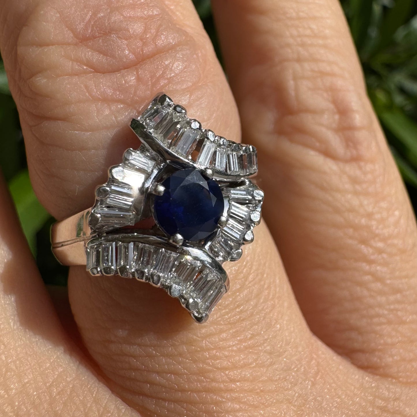 טבעת זהב לבן 14 קראט בעיצוב גלאם עתיק, משובצת ספיר כחולה עגולה במרכז, ומשובצת יהלומים בחיתוך באגט היוצרים צורת מניפה סימטרית ומרקם תלת־ממדי מרהיב.