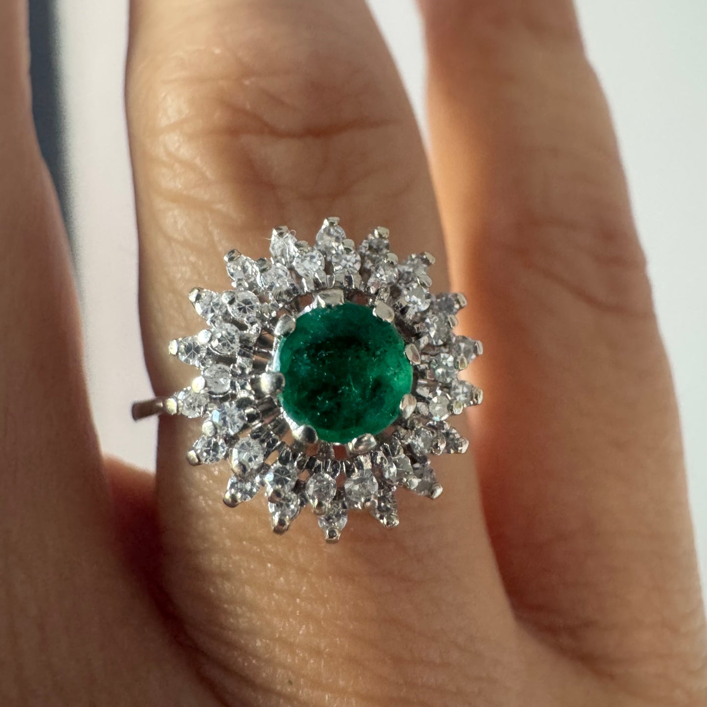 טבעת זהב לבן 14 קראט, משובצת אבן אמרלד ירוקה במרכז, מוקפת הילה עשירה של יהלומים בעיצוב קלאסי ואלגנטי. 