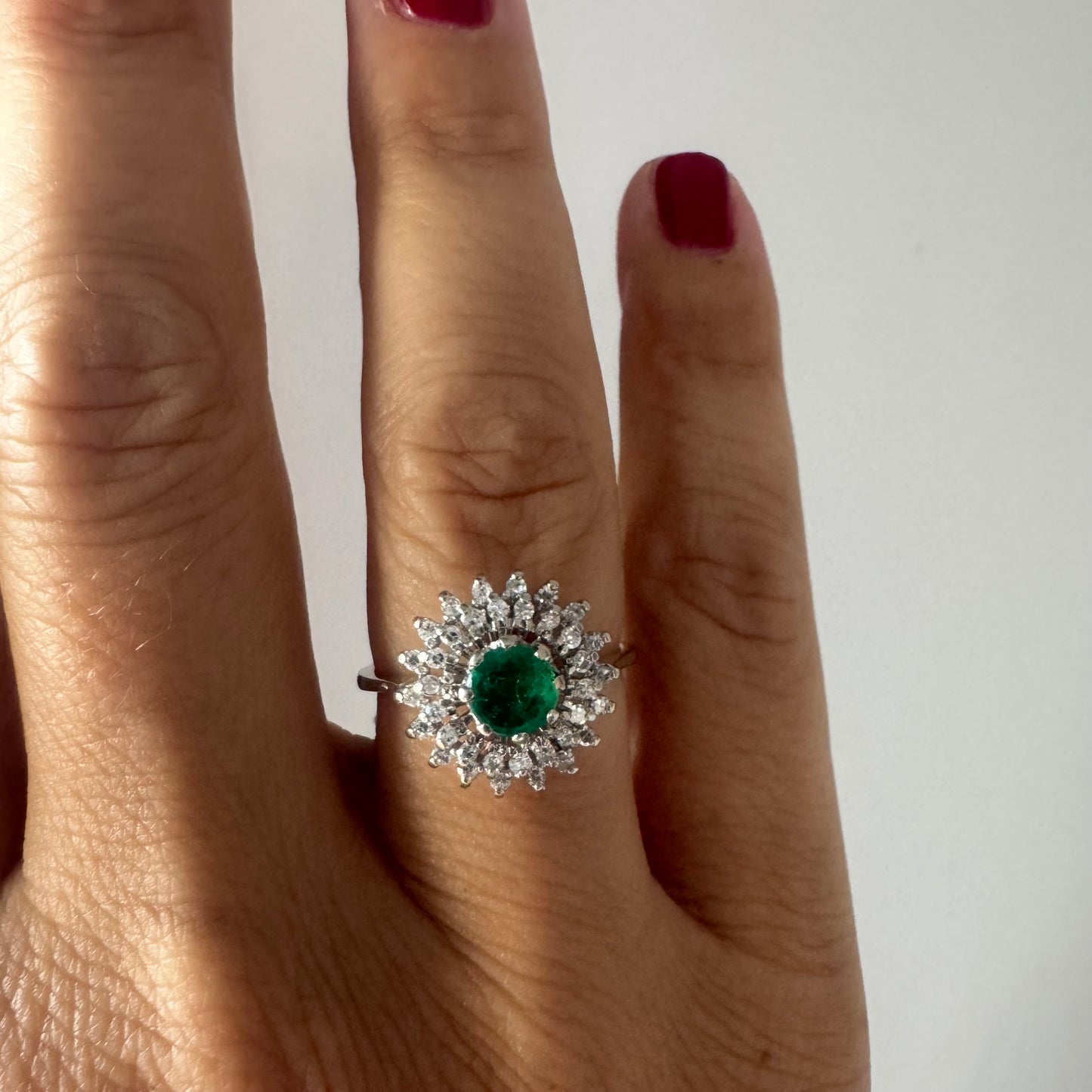 טבעת זהב לבן 14 קראט, משובצת אבן אמרלד ירוקה במרכז, מוקפת הילה עשירה של יהלומים בעיצוב קלאסי ואלגנטי. 