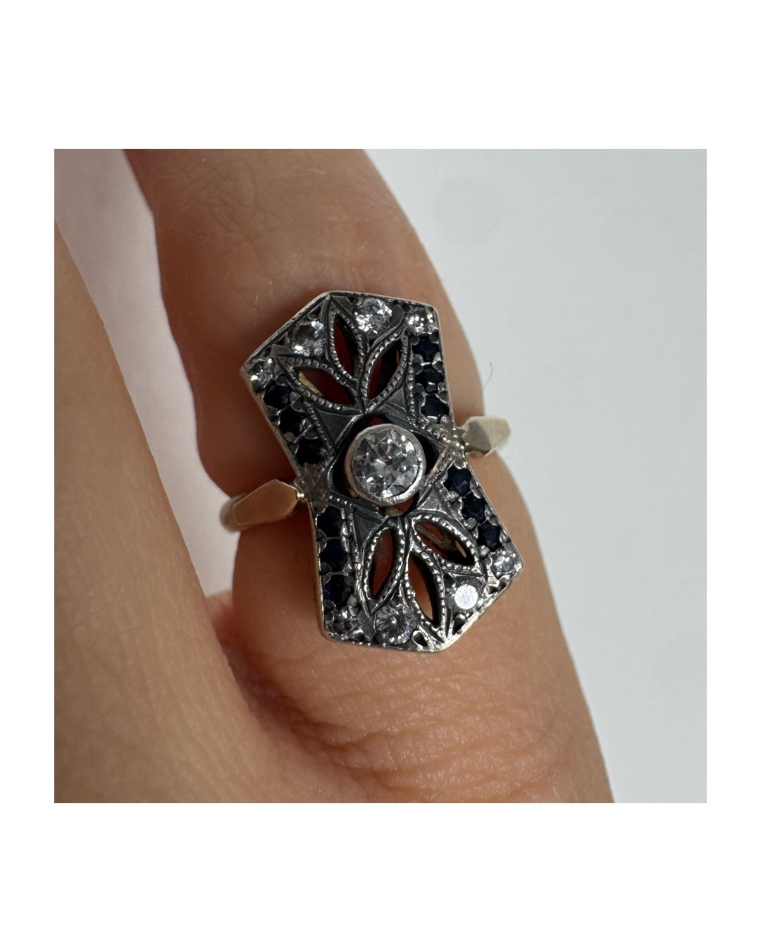 טבעת זהב 14 קראט בעיצוב אר-דקו גיאומטרי, משובצת יהלום מרכזי ומסגרת של אבני ספיר כהות ויהלומים נוצצים.