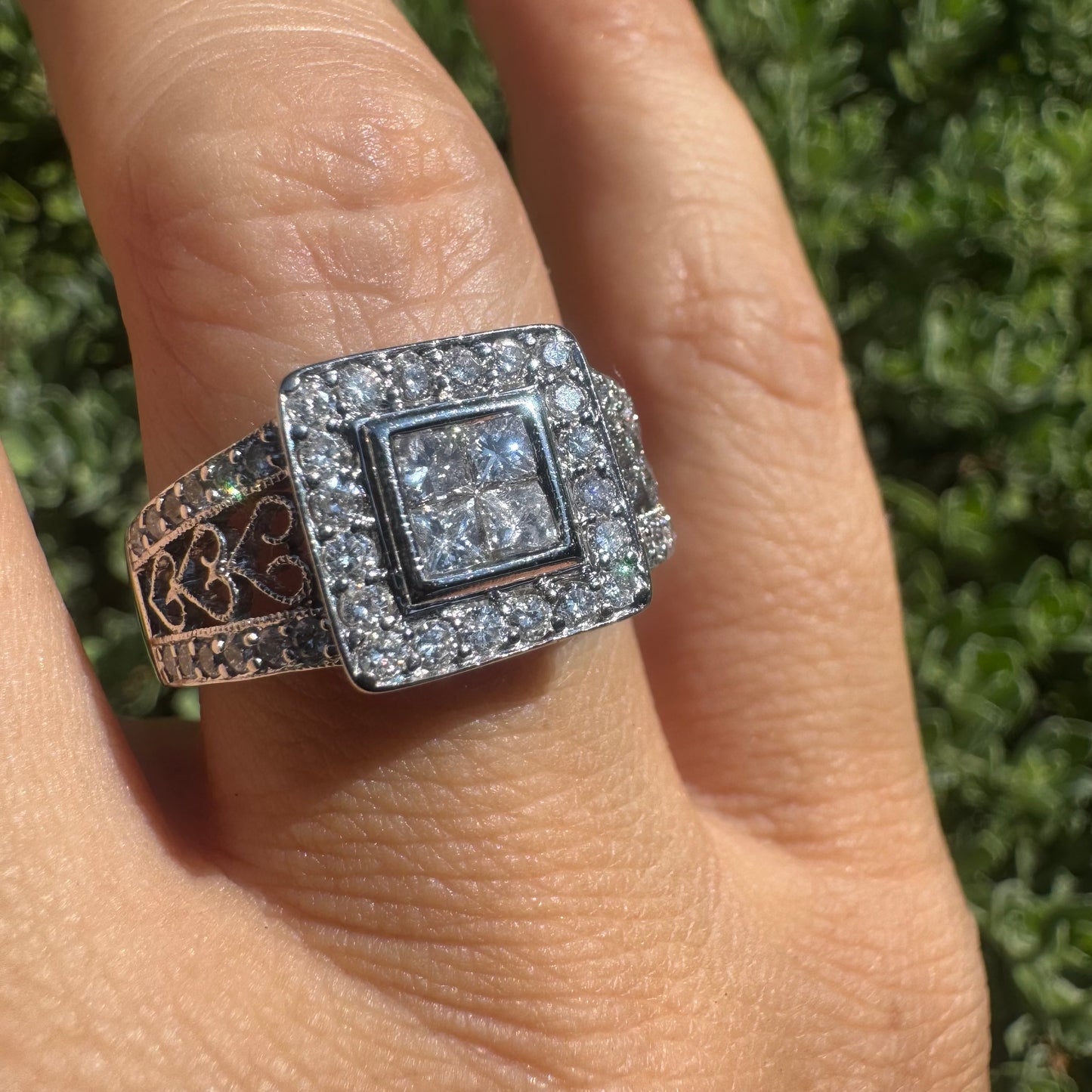 טבעת זהב לבן 14 קראט עם אשכול מרובע של יהלומים בחיתוך פרינסס במרכז, מוקפת בשתי שורות של יהלומים עגולים קטנים, עם עבודת זהב מיוחדת במינה גם על הזרועות