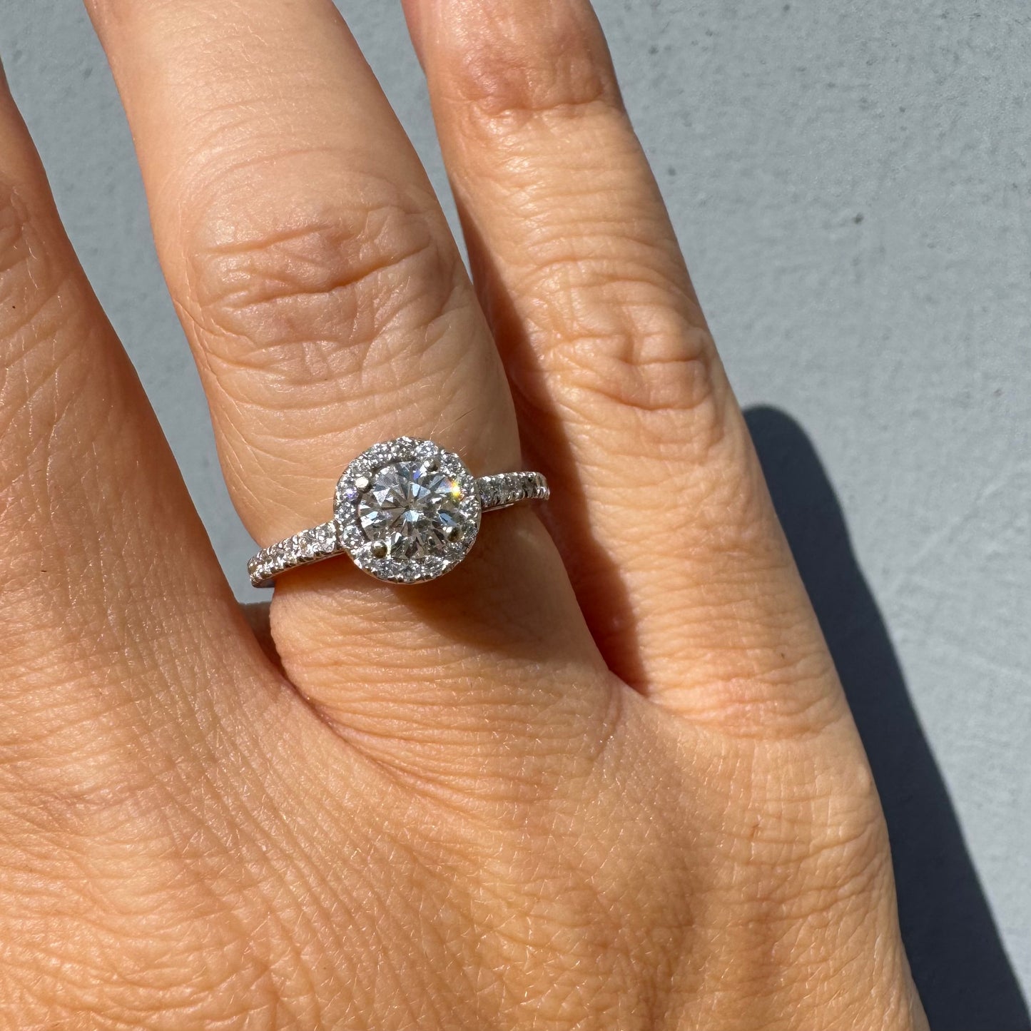 טבעת זהב לבן 14 קראט משובצת יהלום מרכזי עגול והילה נוצצת של יהלומים קטנים, עם יהלומים נוספים על החישוק.