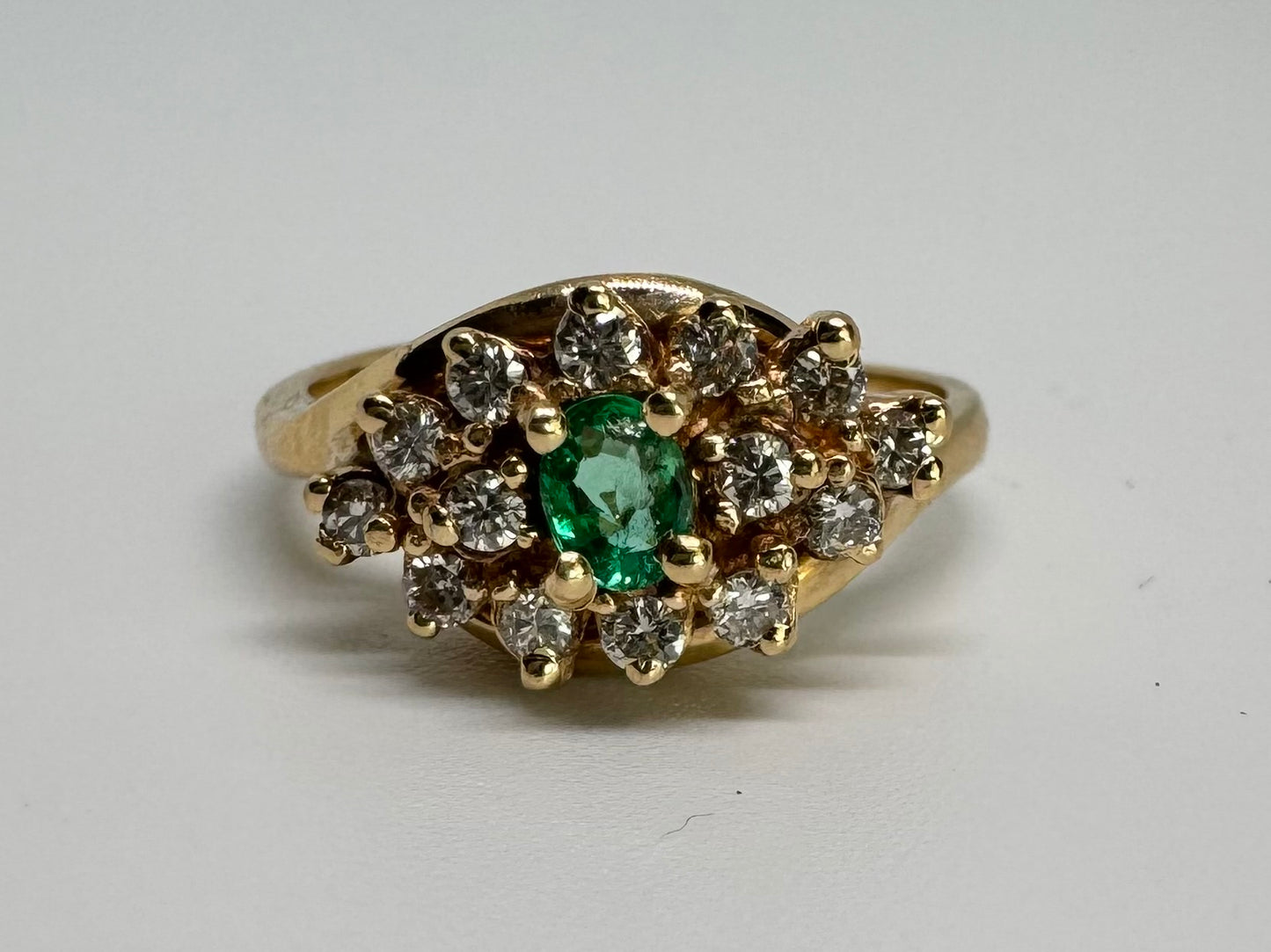 14K gold ring set with Emerald at its center, surrounded by a halo of brilliant-cut diamonds arranged in a floral-inspired cluster