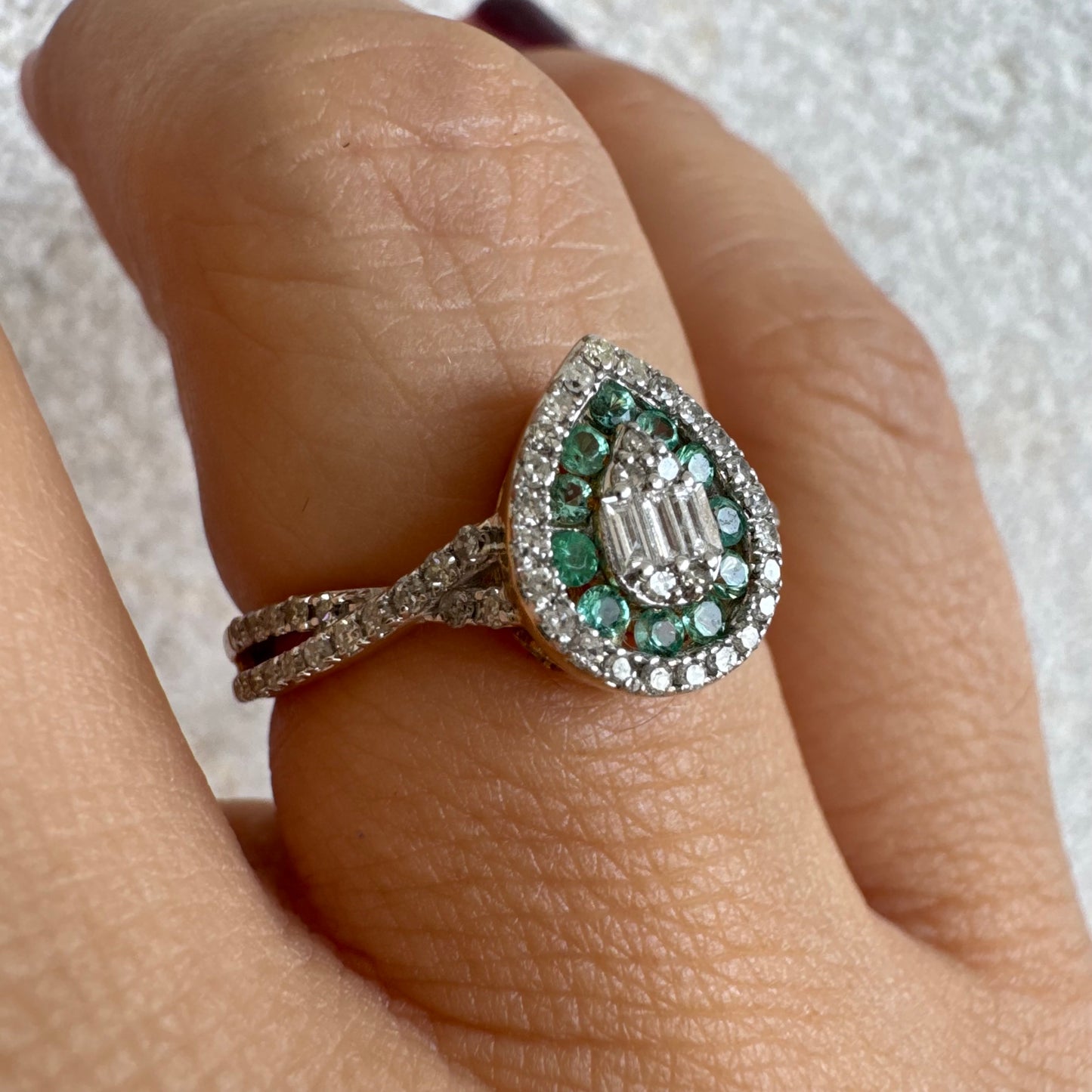 טבעת זהב צהוב 14 קראט בעיצוב טיפה (pear shape), משובצת יהלומים לבנים בשיבוץ הילה כפולה (double halo) ואבני אמרלד קטנות המקיפות מרכז ייחודי של יהלומים בחיתוך באגט