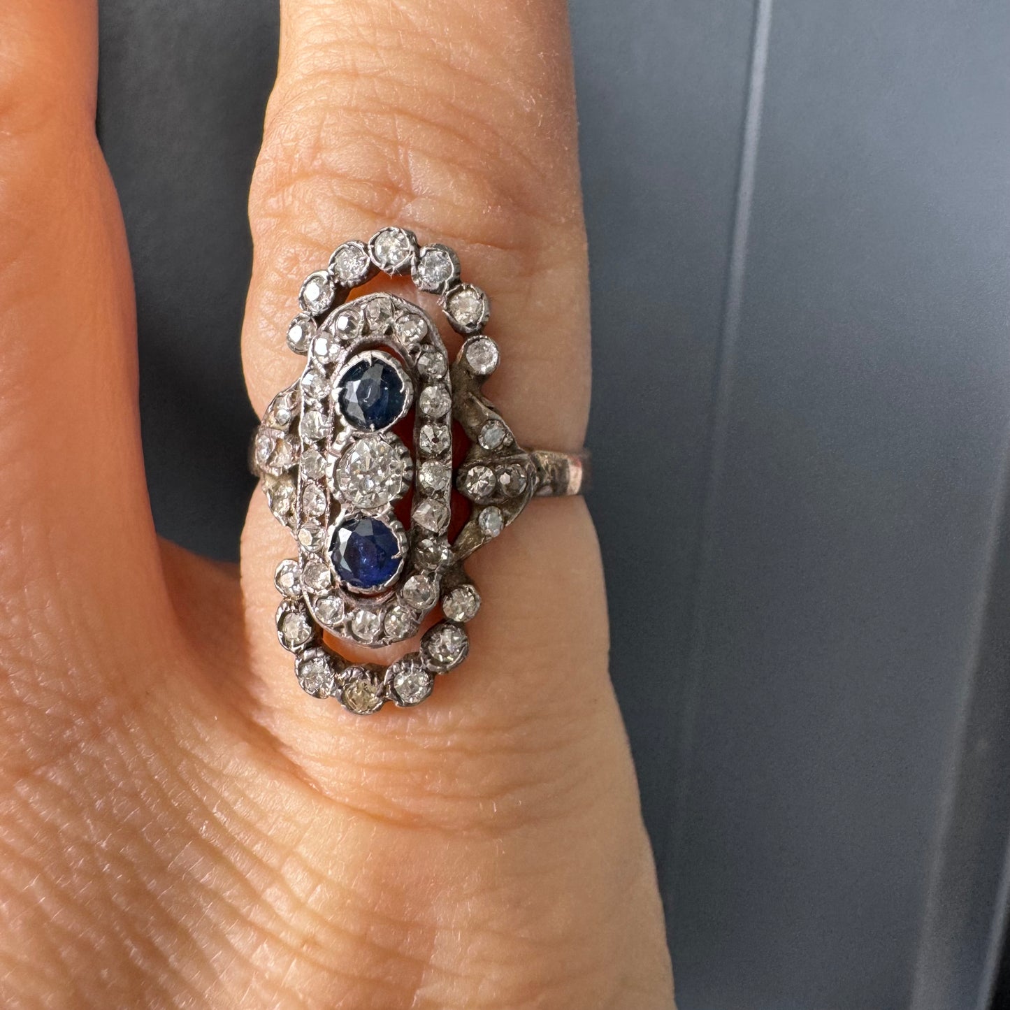 טבעת זהב לבן 14 קראט עתיקה עוצרת נשימה, בעיצוב מאורך ואלגנטי, המשובצת בשלישיית אבנים — שתי אבני ספיר בצבע כחול עמוק ובאמצע משובץ יהלום מרכזי בחיתוך עתיק.