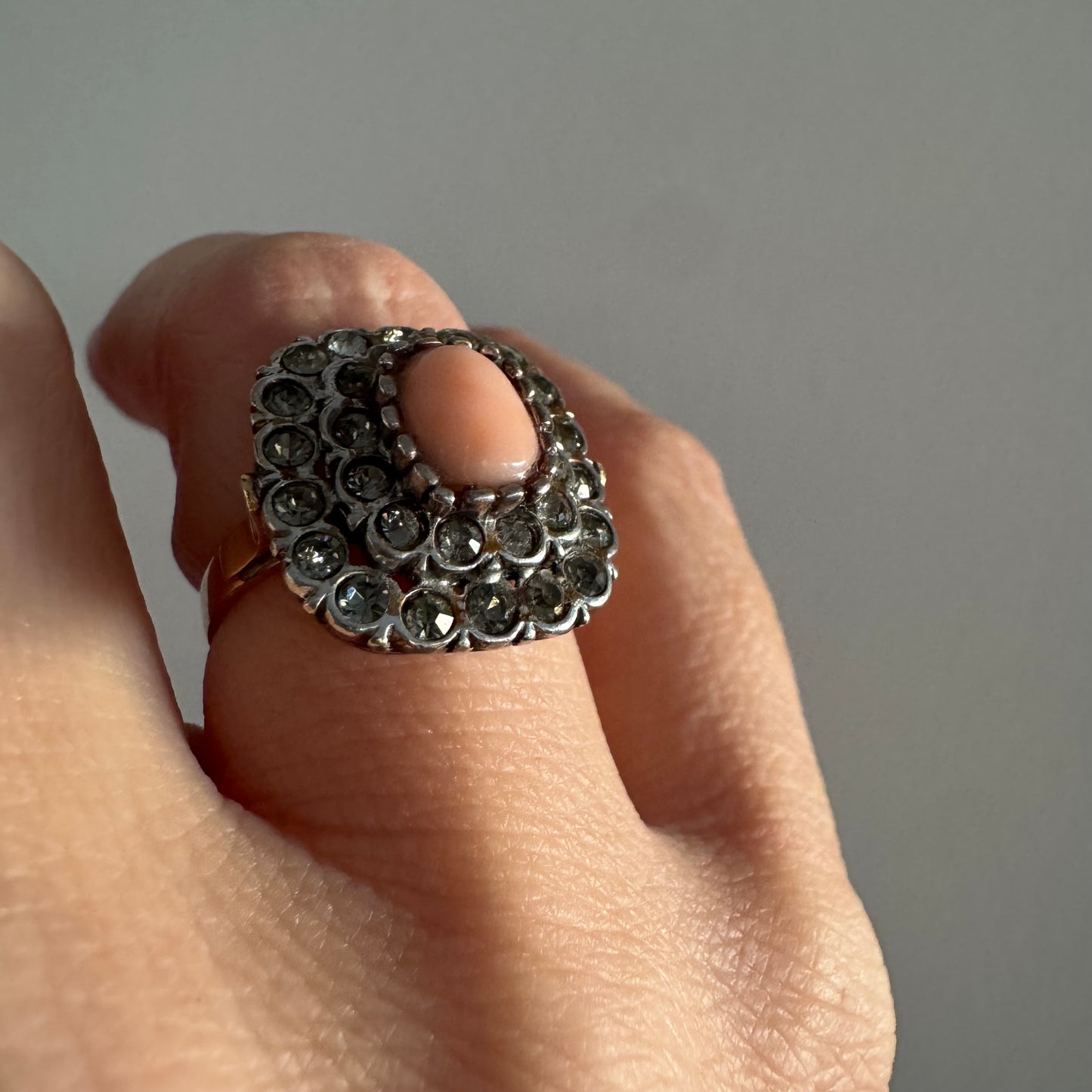 טבעת וינטג׳ עם חלק תחתון מזהב 14 קראט וחלק עליון עשוי כסף משובצת אבן קורל בהירה במרכז ומסביבה אבנים בהירות שאינן יהלומים.